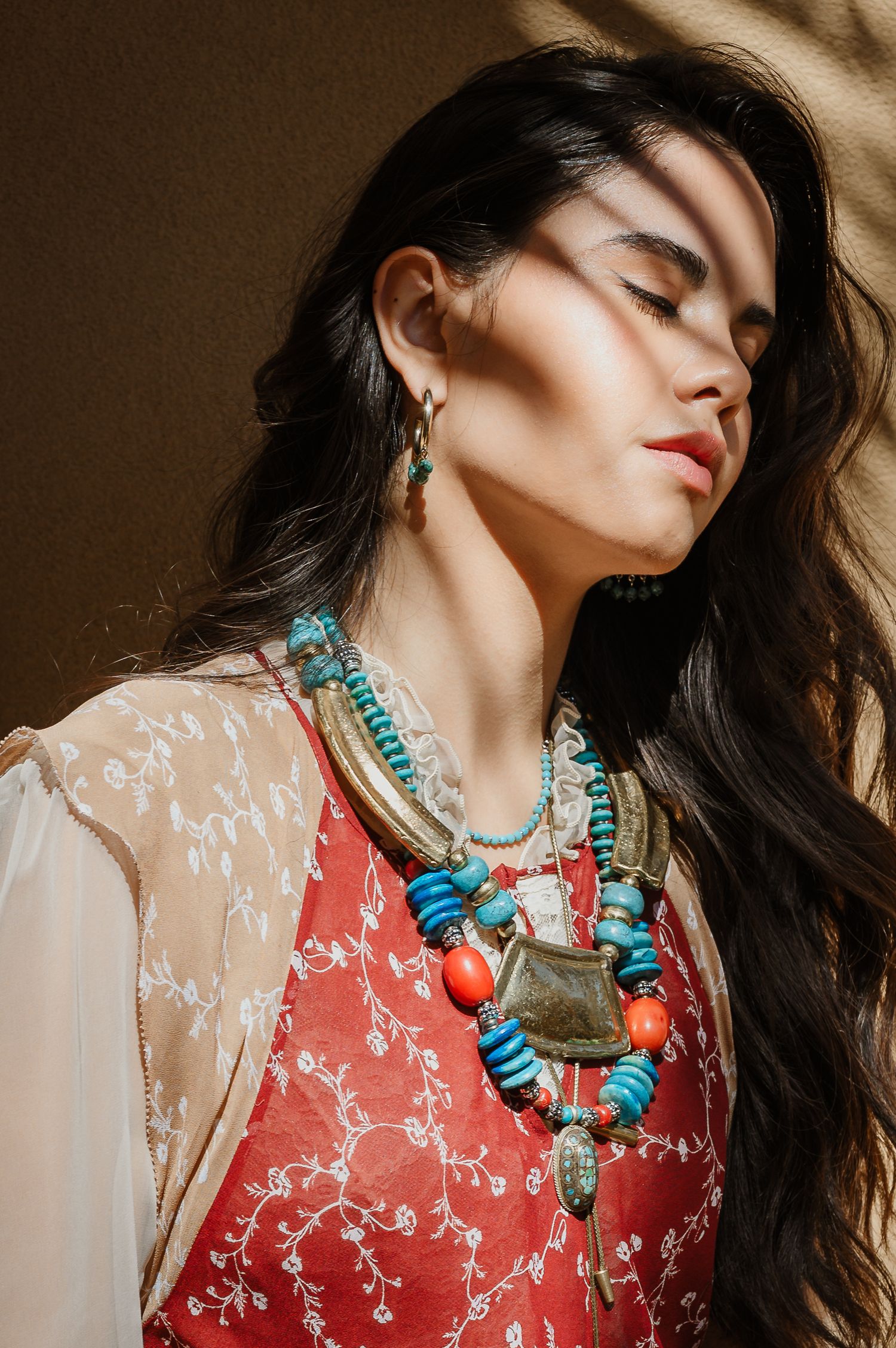 Woman wearing turquoise and coral beaded necklace with ethnic pendant, red embroidered dress, in warm sunlight.