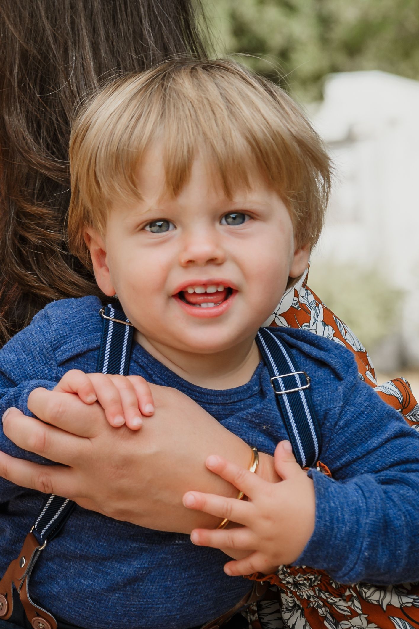 Smiling toddler with blonde hair and blue eyes in a blue shirt, held by an adult outdoors.