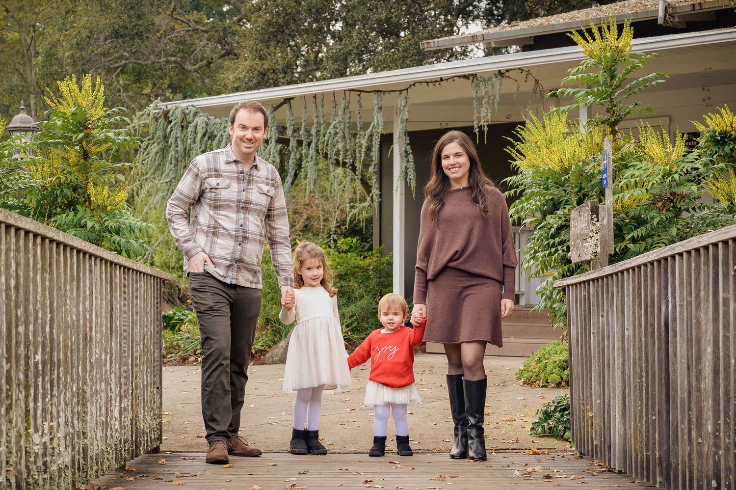 Family of four posing on a garden path, parents holding hands with two young children outside a charming home.