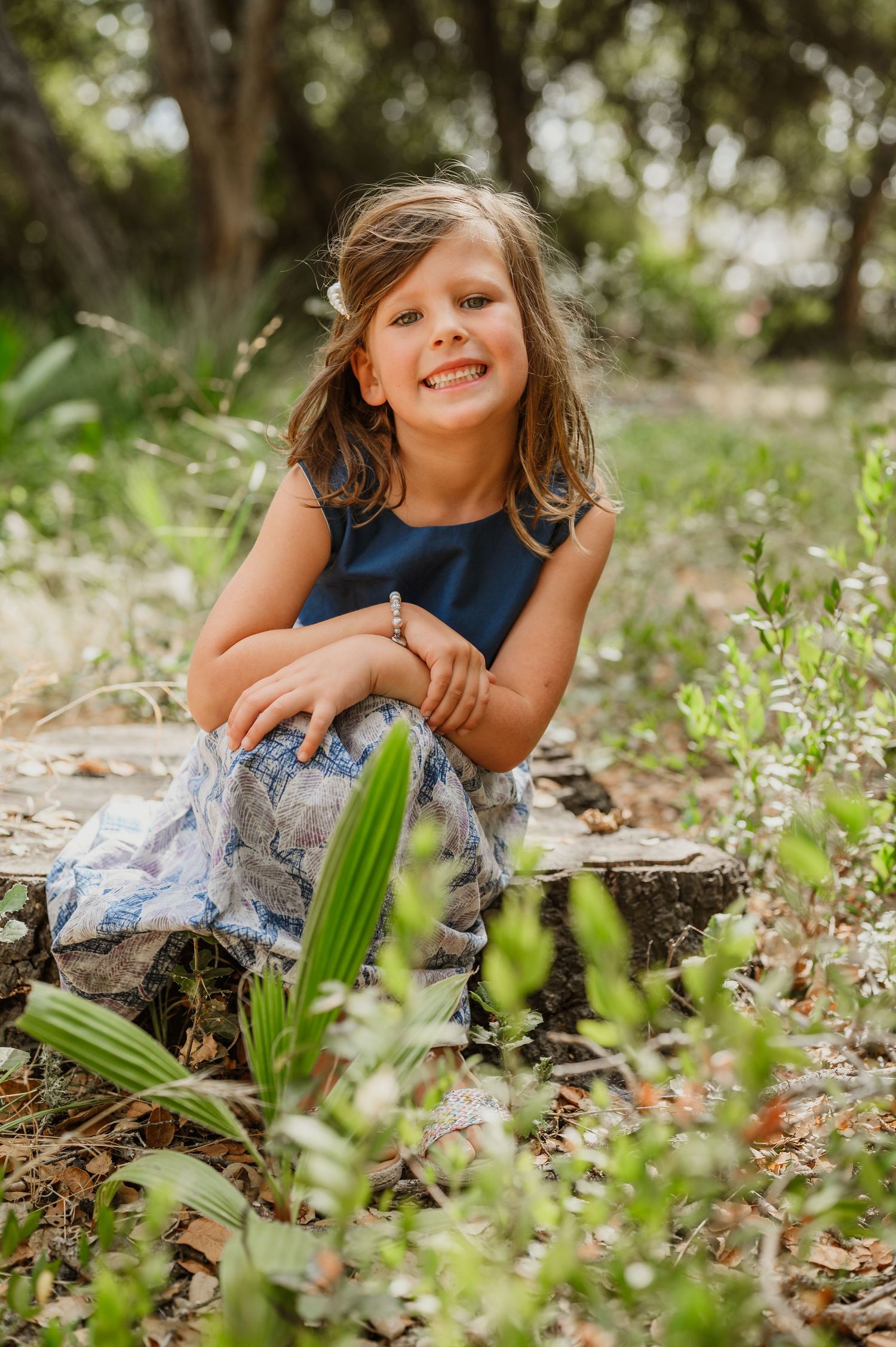 A smiling young girl in a navy top and floral skirt sits outdoors among green plants and wildflowers in a natural setting.
