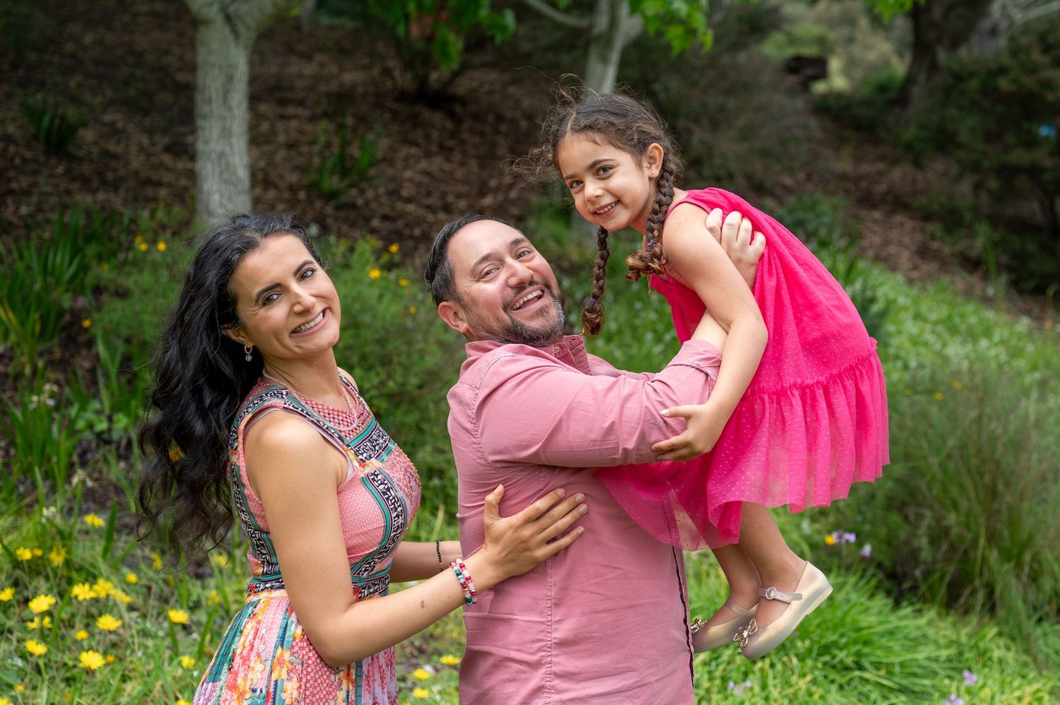 Happy family portrait with parents and young daughter in pink dress outdoors in lush green garden setting.