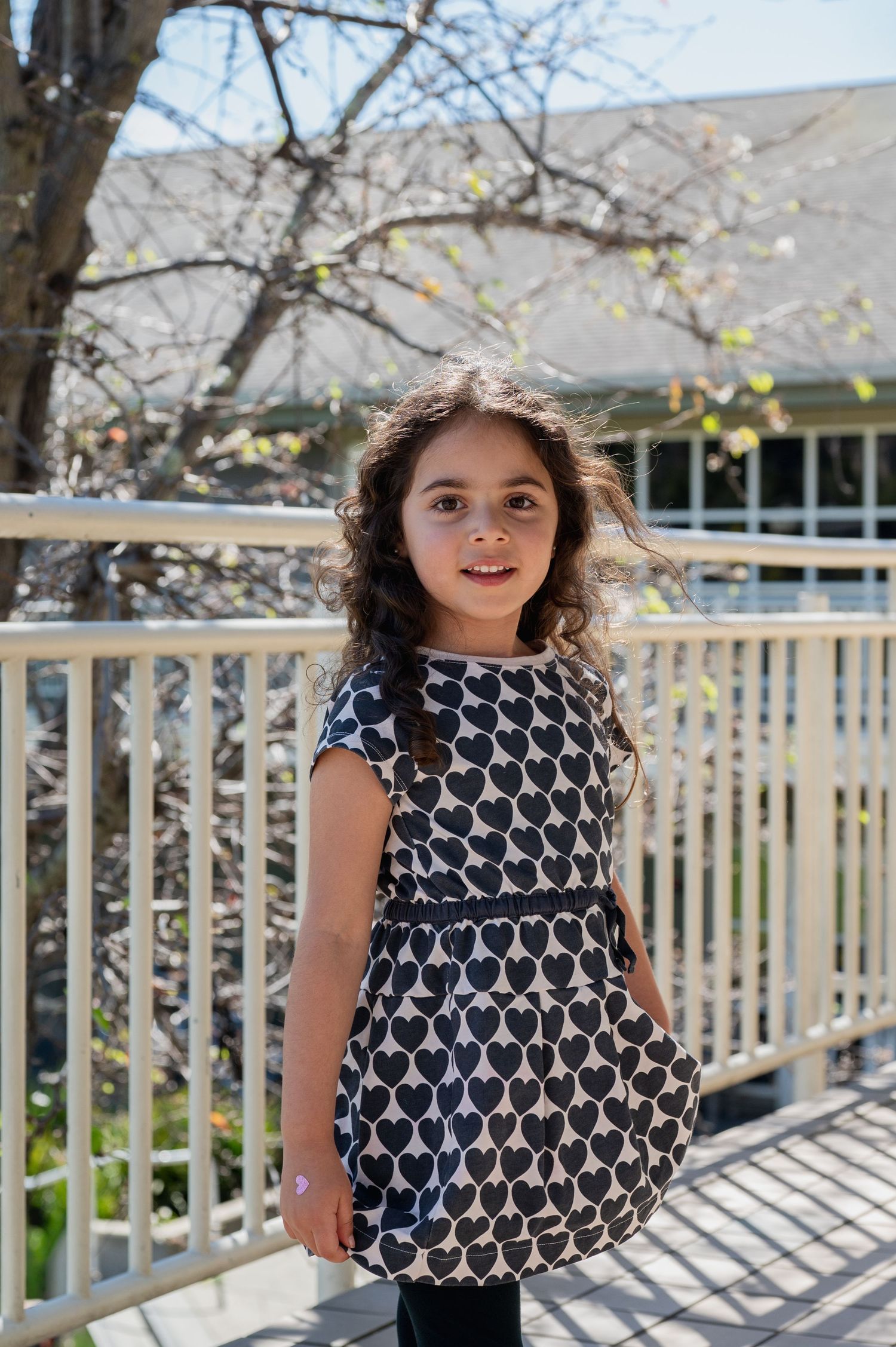 Young girl in heart-print dress standing on sunny outdoor bridge with bare trees.