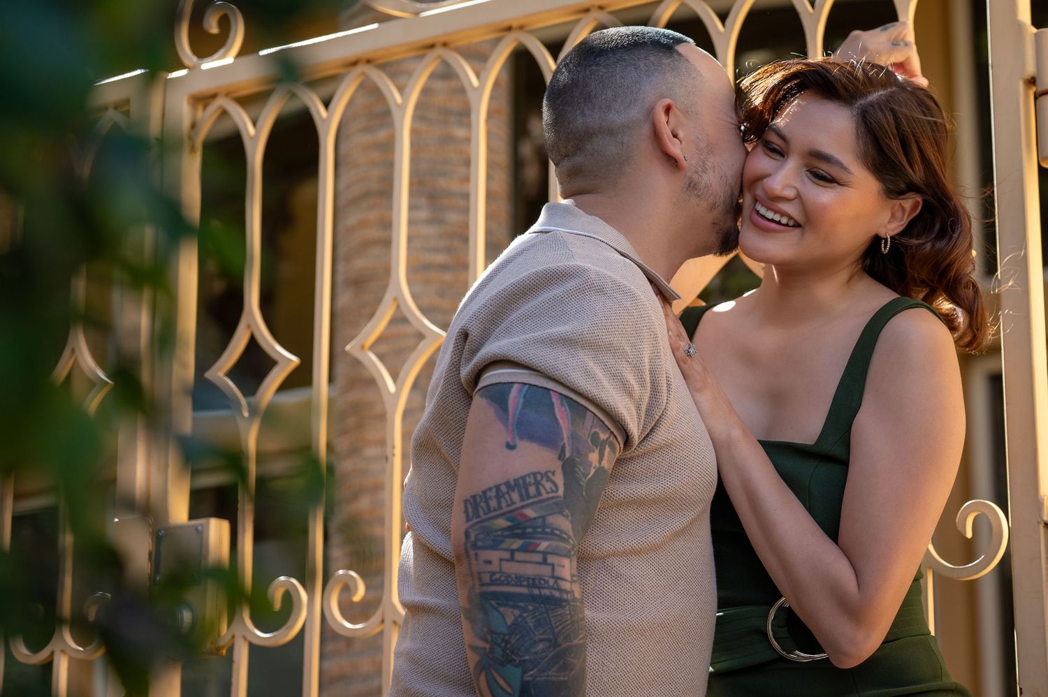 A tattooed man kisses a smiling woman in a green dress near ornate golden gates in warm sunlight.