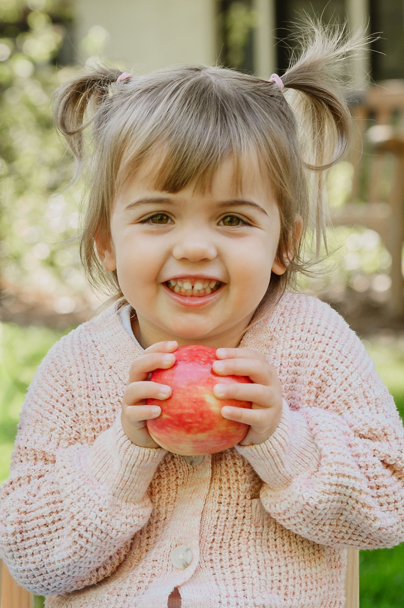 Smiling toddler girl with pigtails holding a red apple outdoors in a pink knit cardigan.