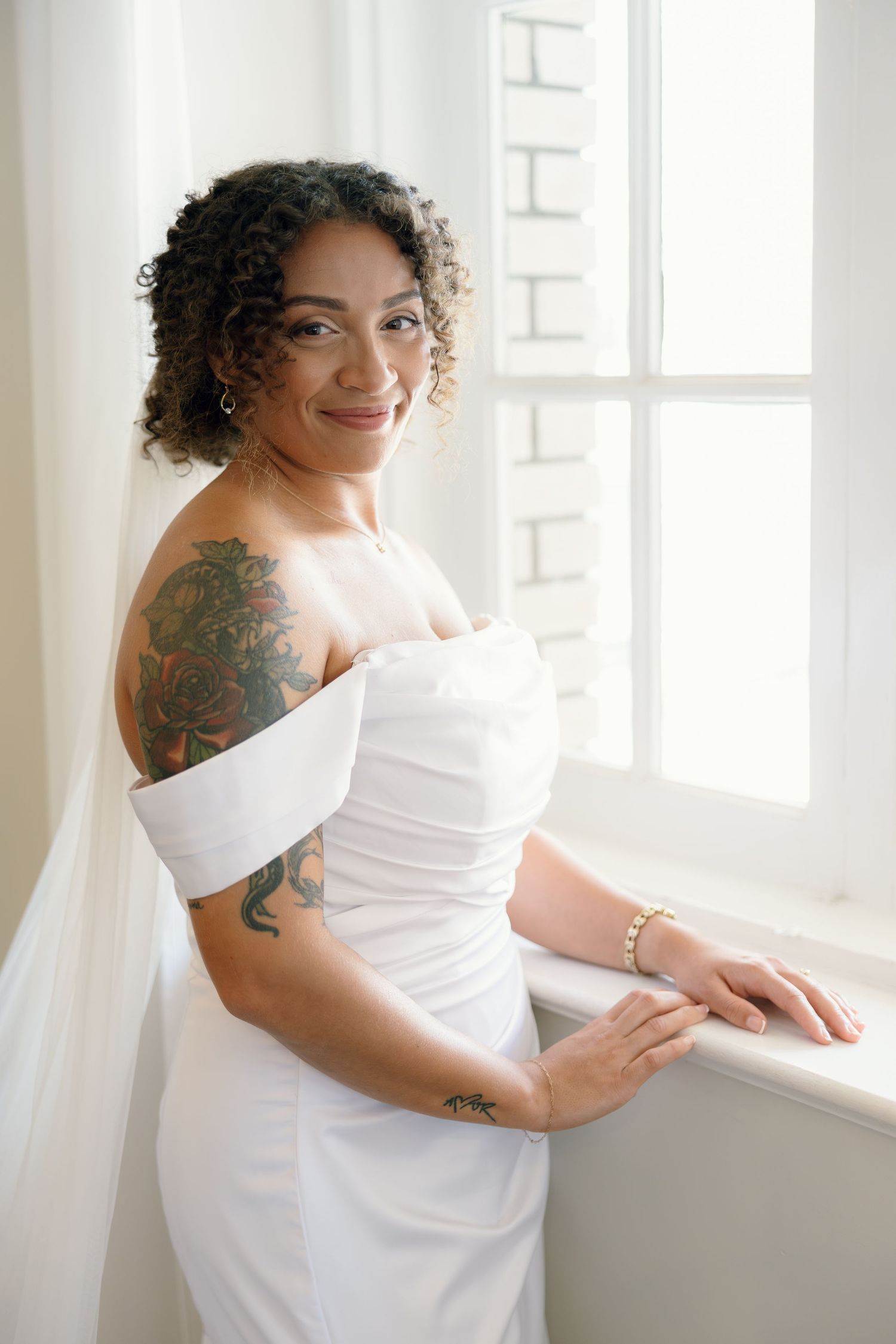 Smiling bride in white off-shoulder gown with floral tattoos poses by a bright window wearing a veil.