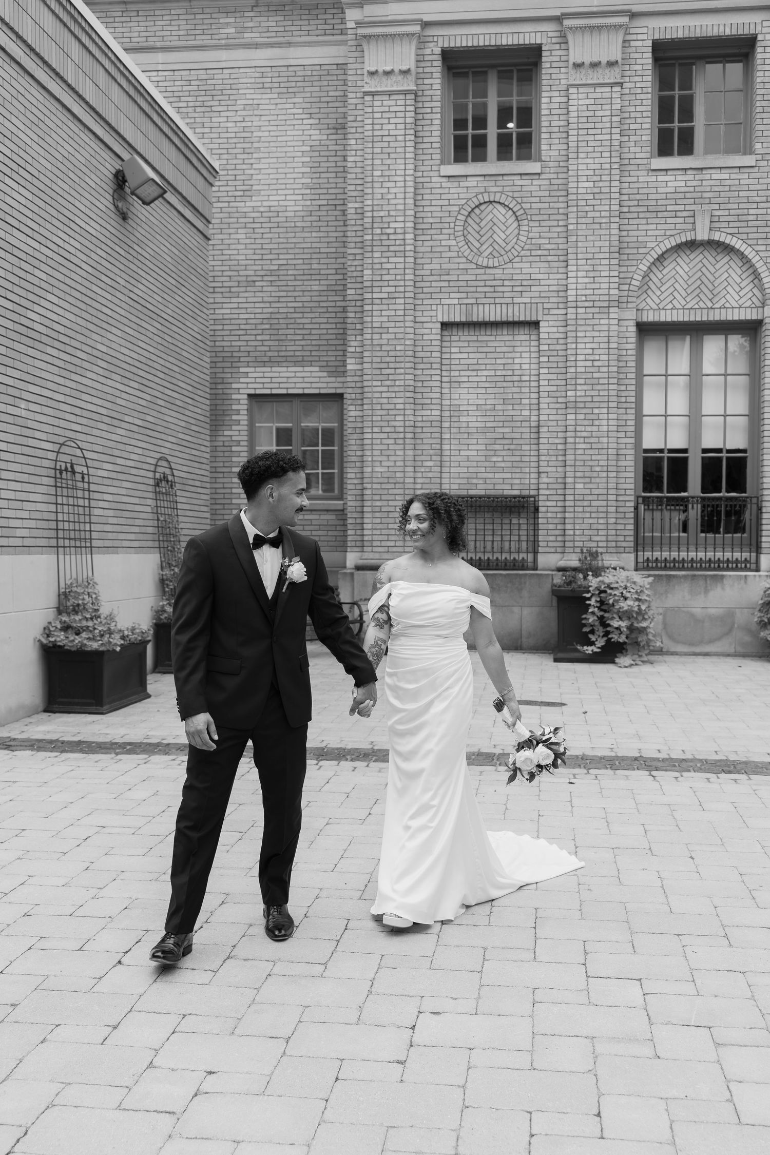 Black and white photo of a bride and groom holding hands, walking on cobblestone outside a brick building.