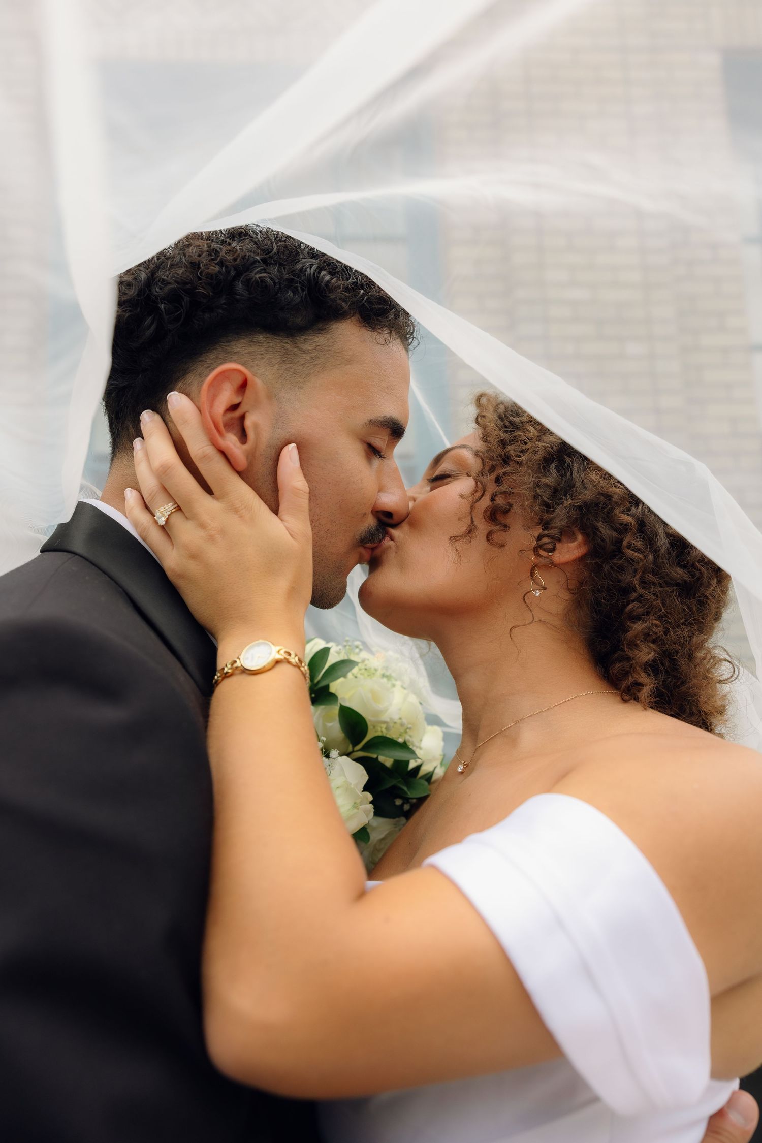 A bride and groom share a romantic kiss under a flowing white veil, bride holding white roses bouquet.