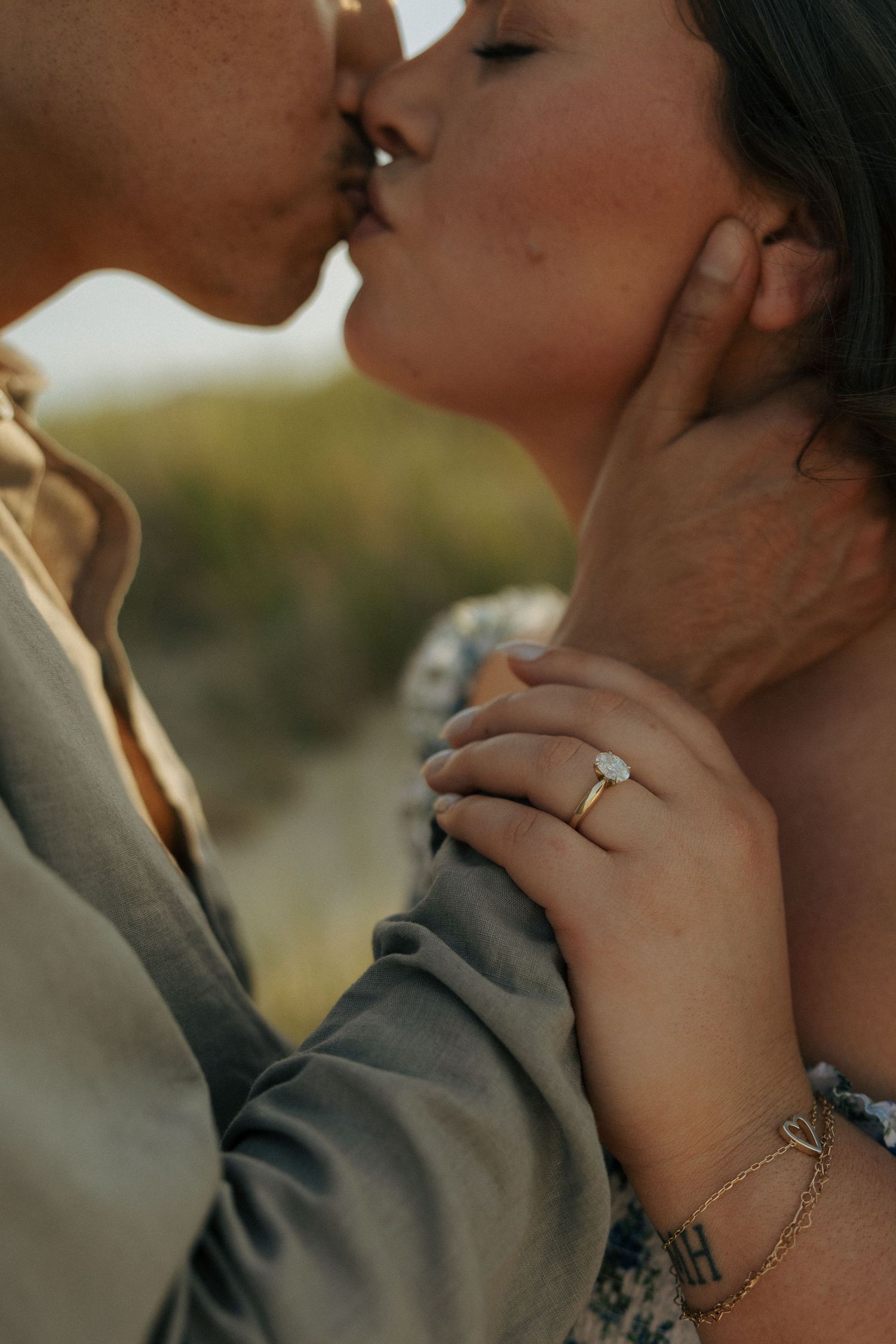 Close-up of couple kissing during engagement session, woman displaying diamond solitaire ring on gold band.