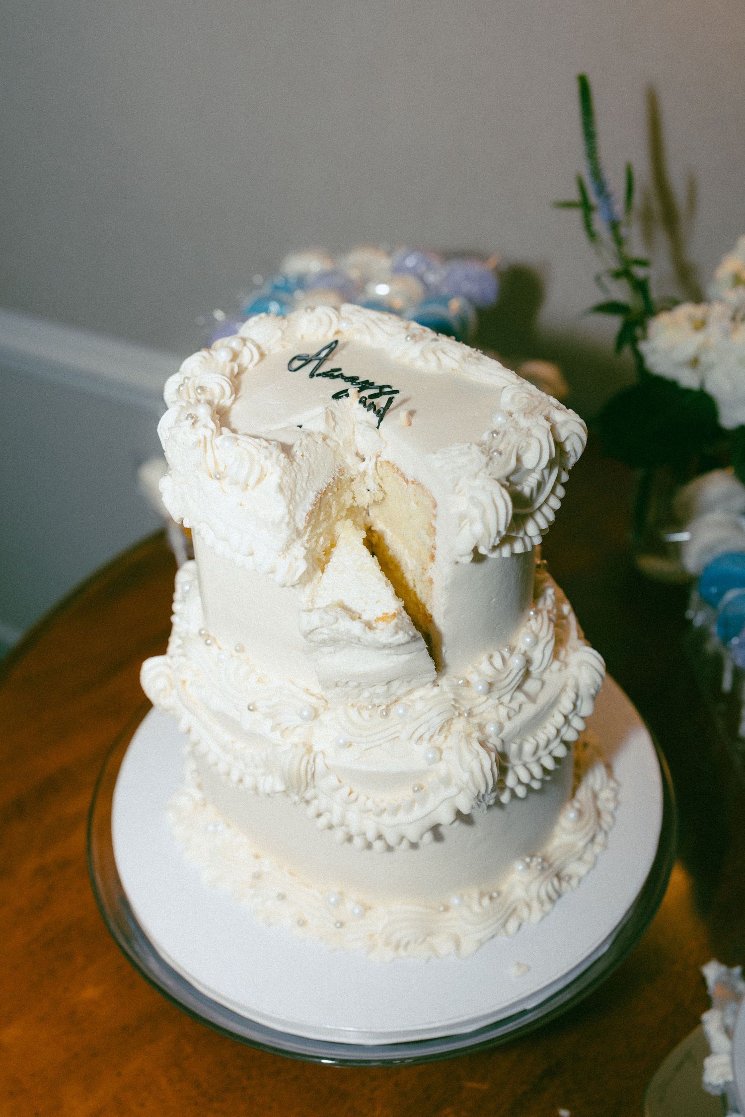 Two-tier white wedding cake with ornate frosting details and a script topper, partially sliced to reveal yellow interior.