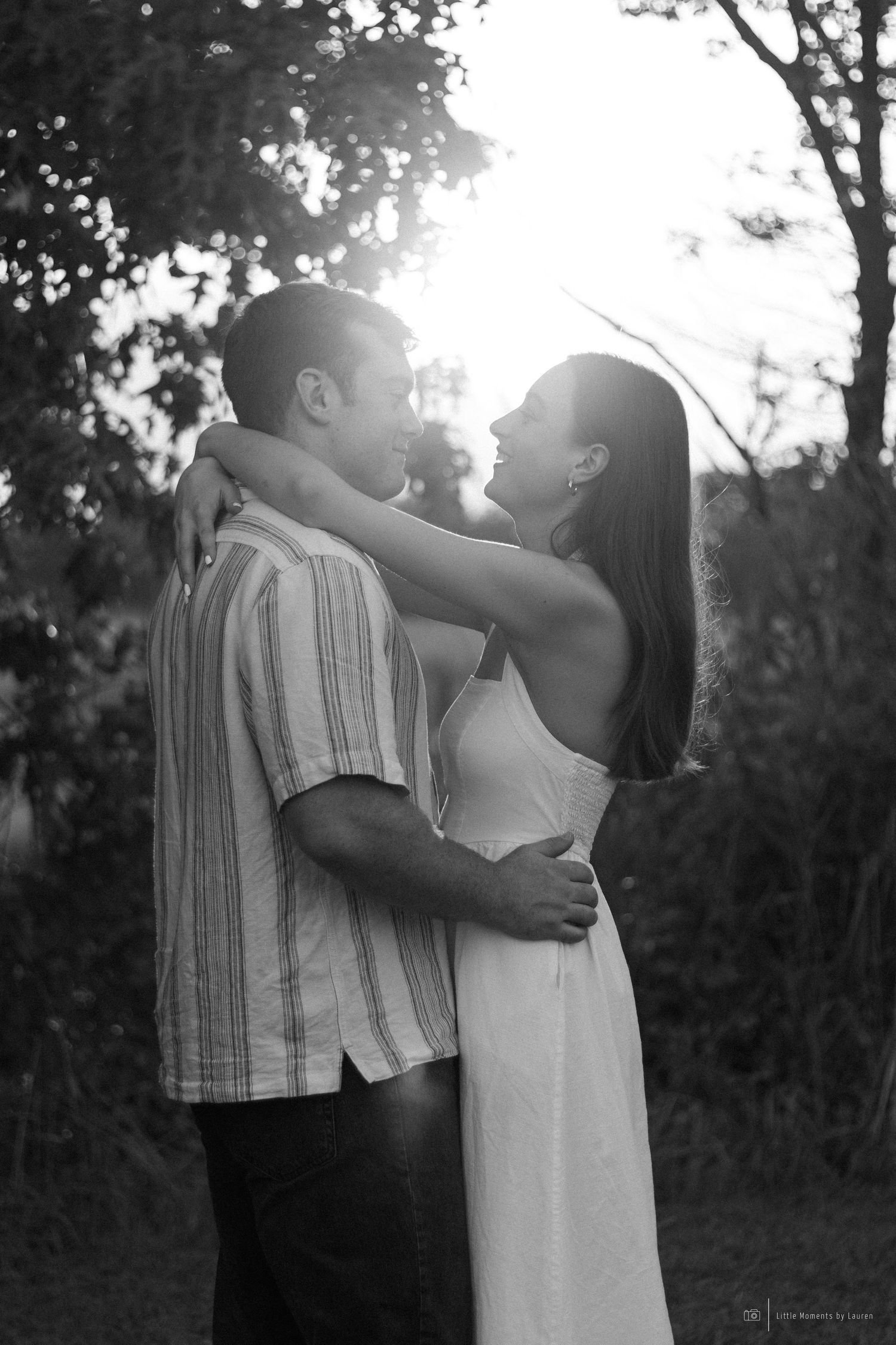 Black and white photo of a couple embracing at sunset in a lush outdoor setting.