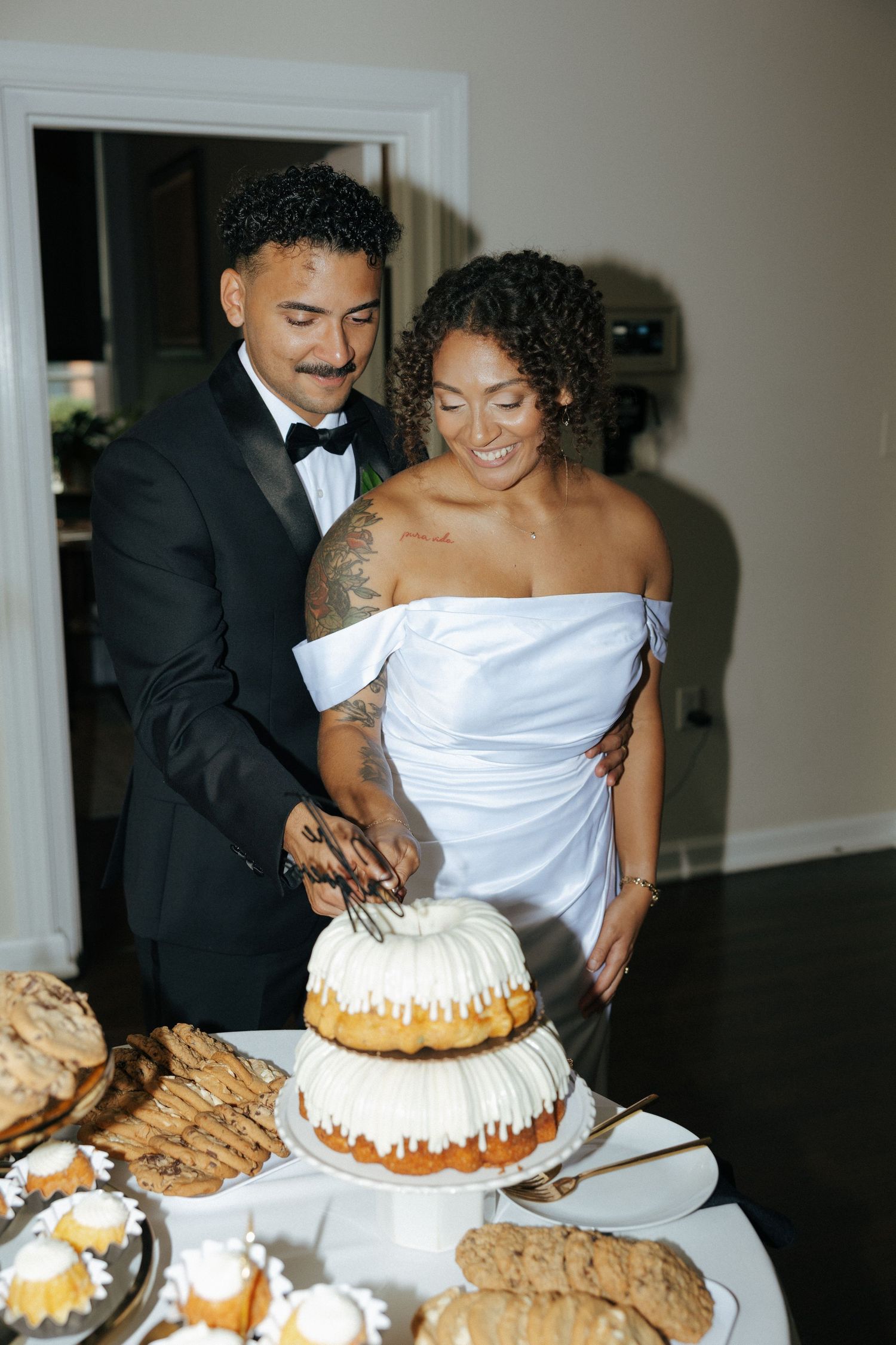 Newlywed couple cutting their white bundt wedding cake, groom in tuxedo and bride in off-shoulder gown.