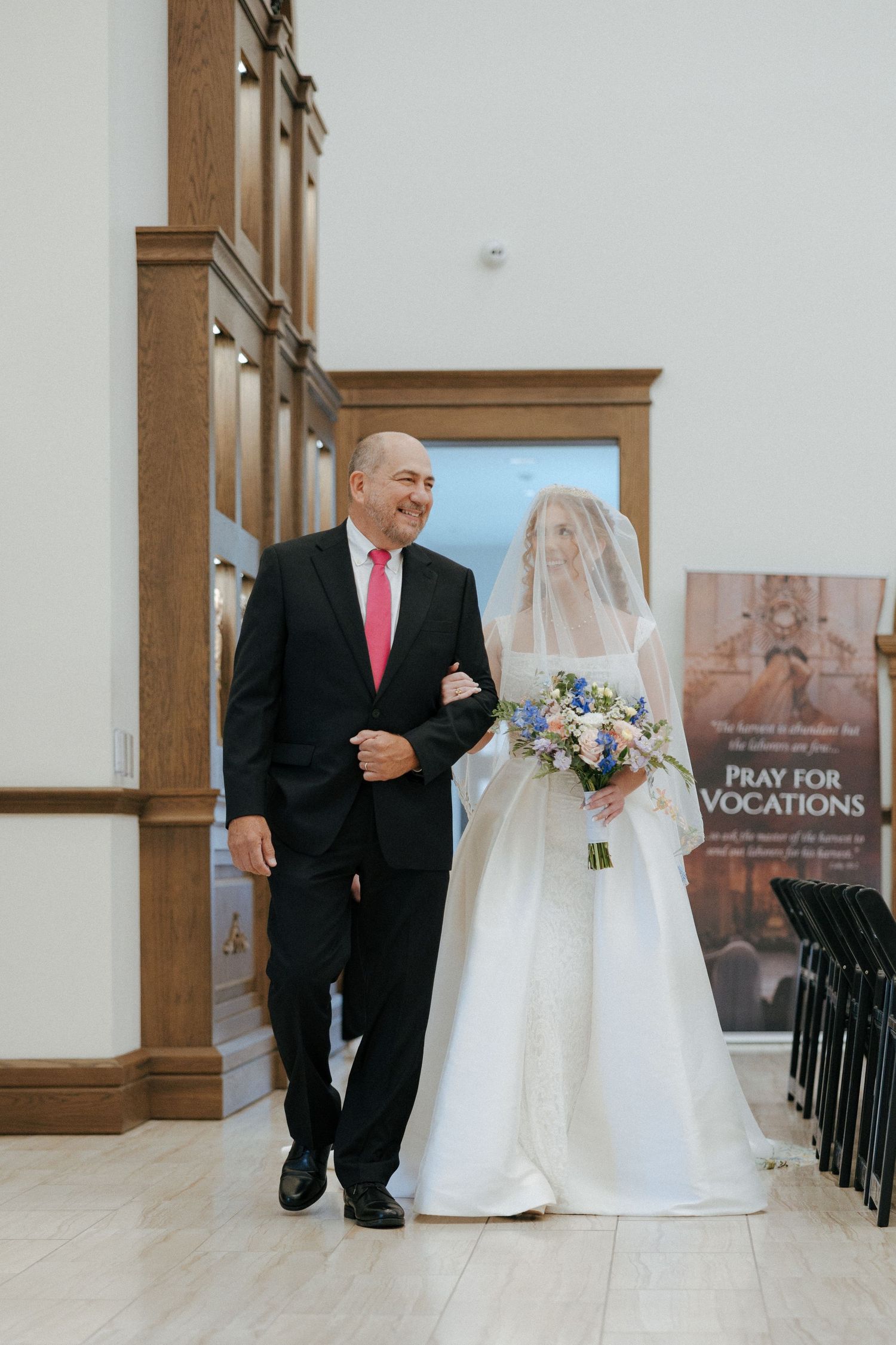 Father escorts bride in white gown and veil down church aisle, holding blue and white bouquet at wedding ceremony.
