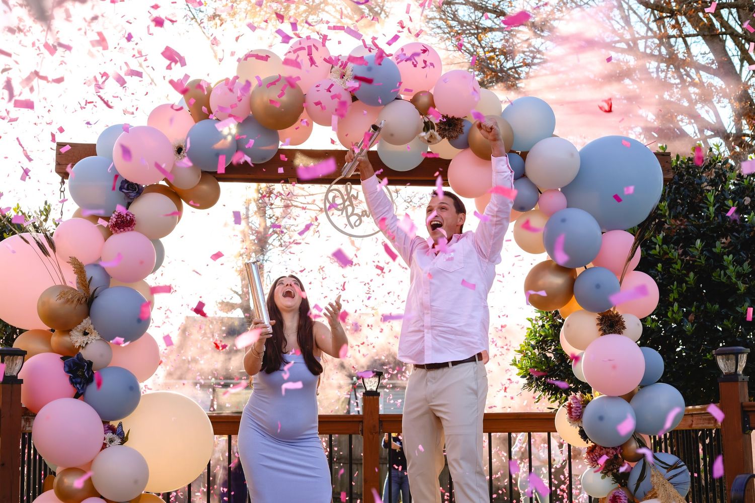 Couple celebrating gender reveal with pink confetti under colorful balloon arch outdoors.