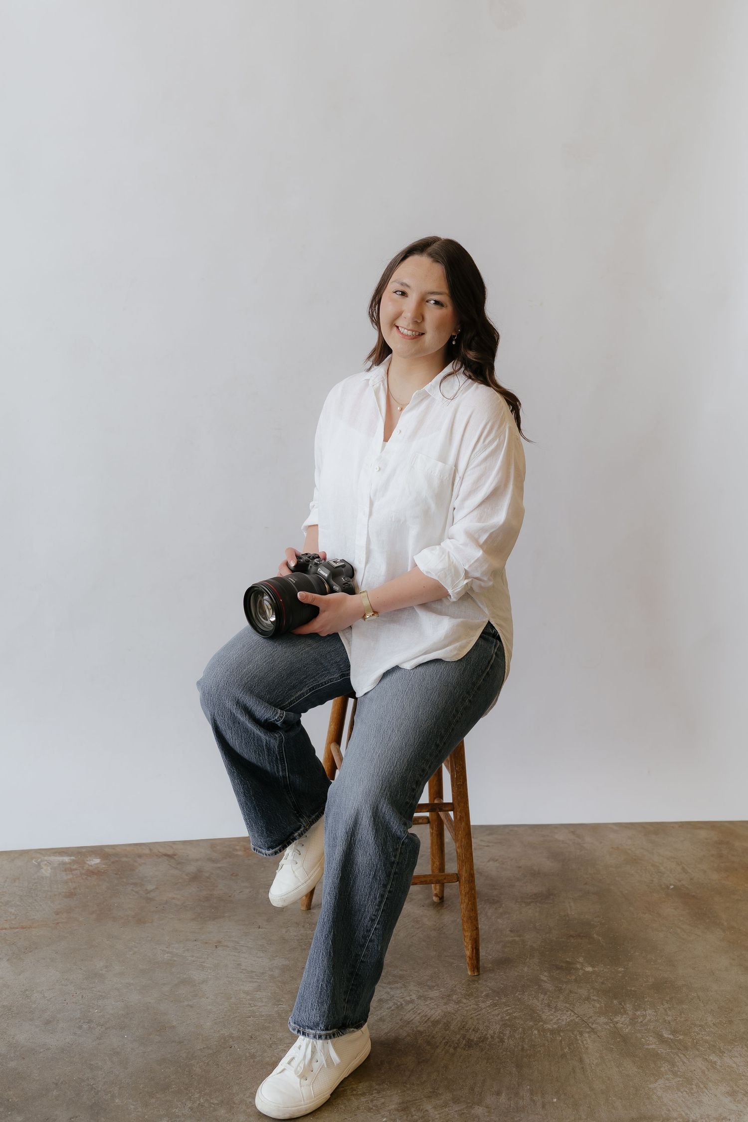 Professional wedding photographer smiling while seated on a stool, holding a camera in a bright studio.