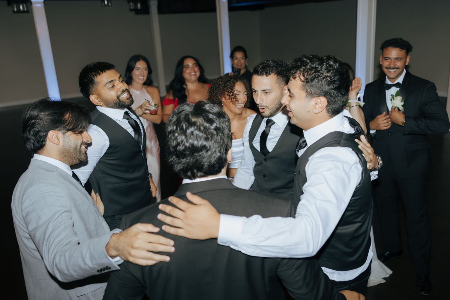 Groomsmen in black vests celebrate together on the dance floor at a wedding reception.