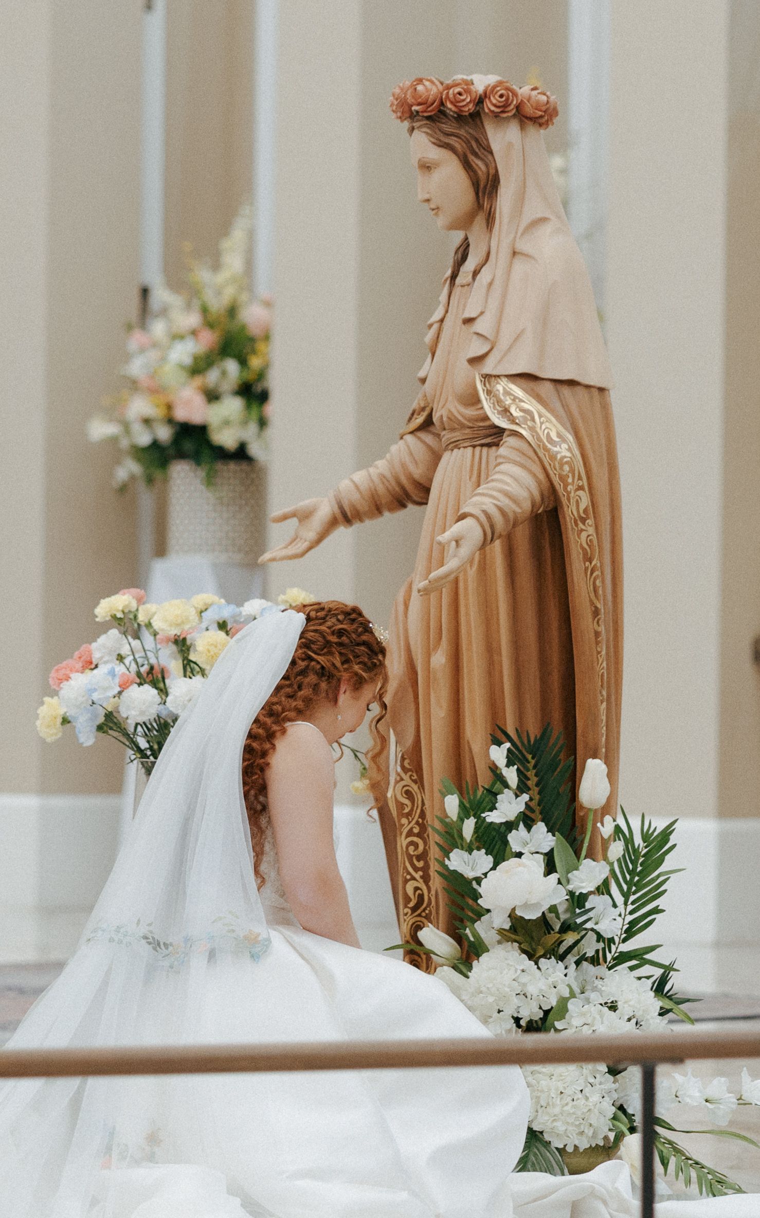 Young girl in white veil kneels before Virgin Mary statue surrounded by flowers during First Communion ceremony.
