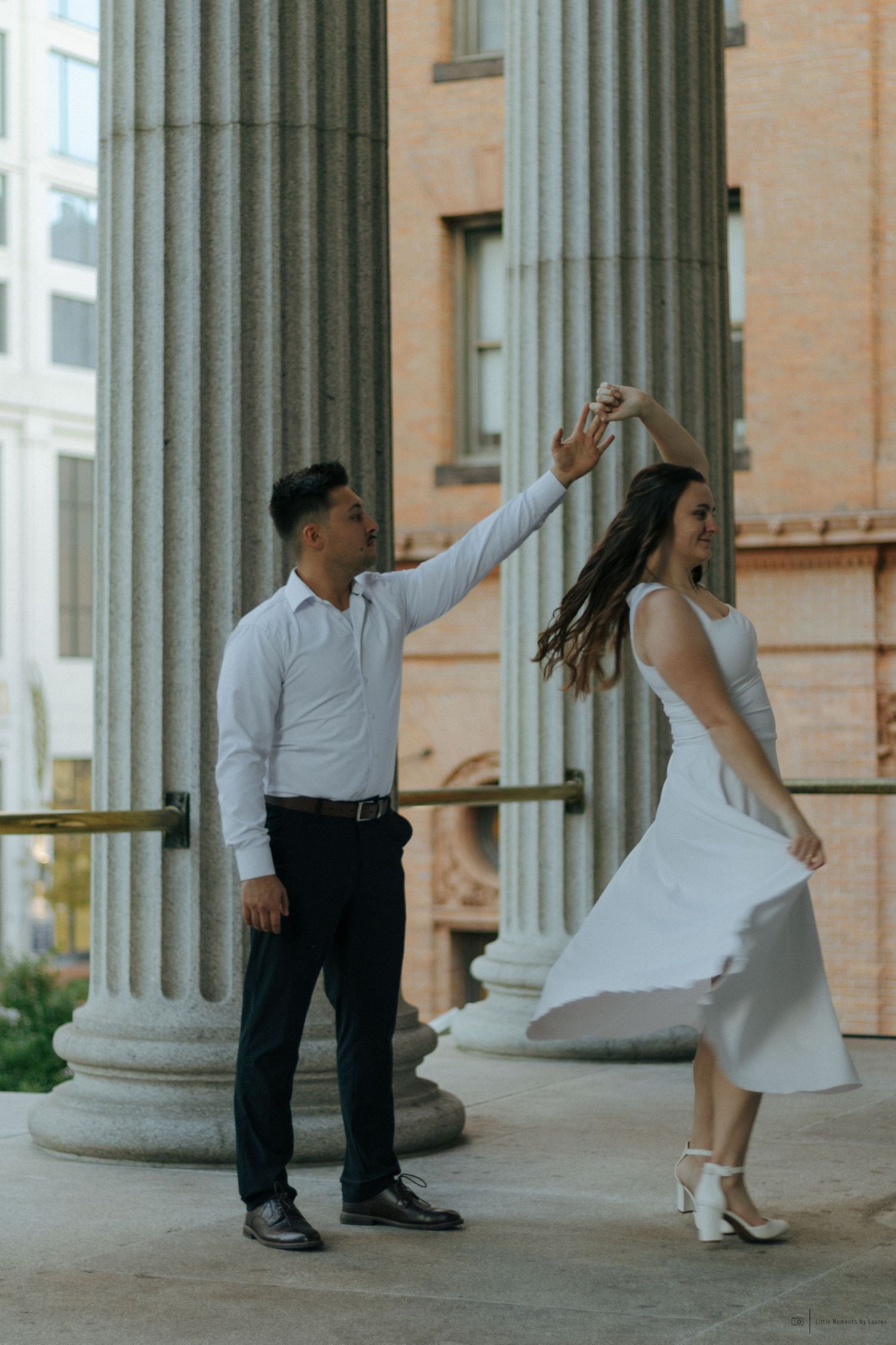 A couple dances outdoors near stone columns, the woman in a white dress spinning gracefully while the man leads her.