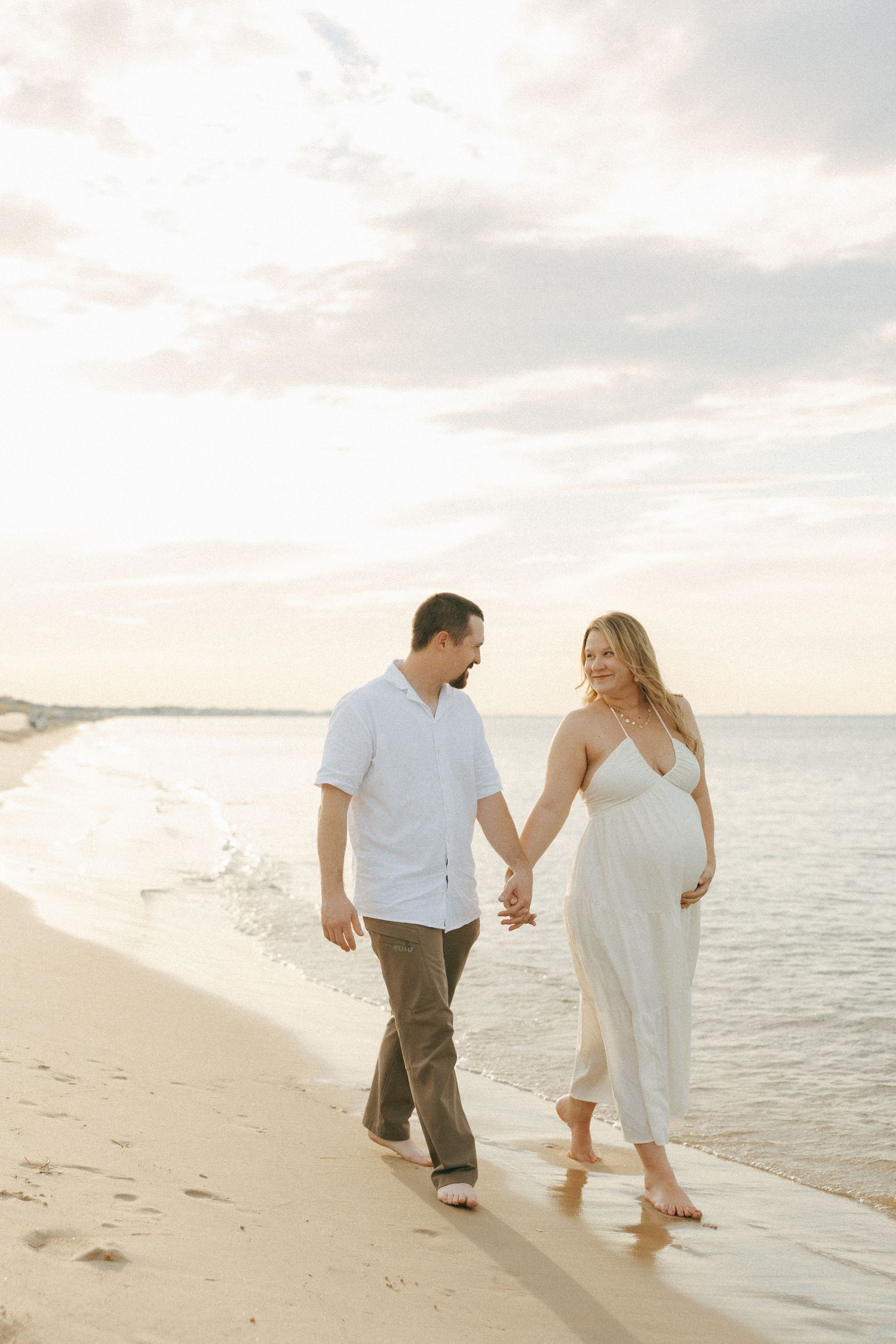 Pregnant couple holding hands walking barefoot on sandy beach at sunset, woman in white dress cradling baby bump.