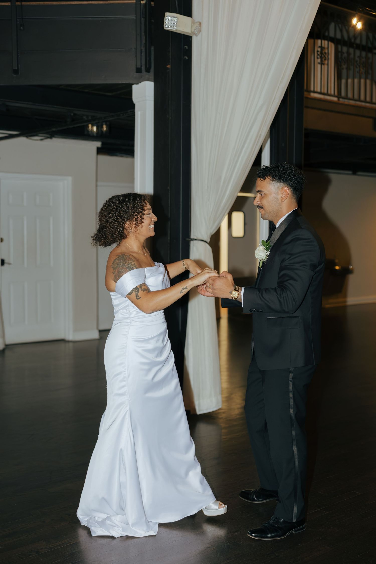 Bride and groom share their first dance in an elegant venue, bride in white off-shoulder gown, groom in black tuxedo.
