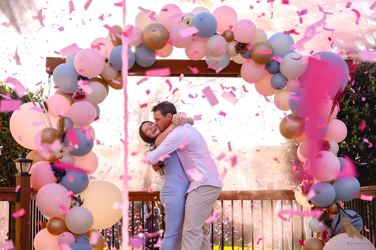 Couple embracing under a colorful balloon arch with pink confetti at a gender reveal party celebration.