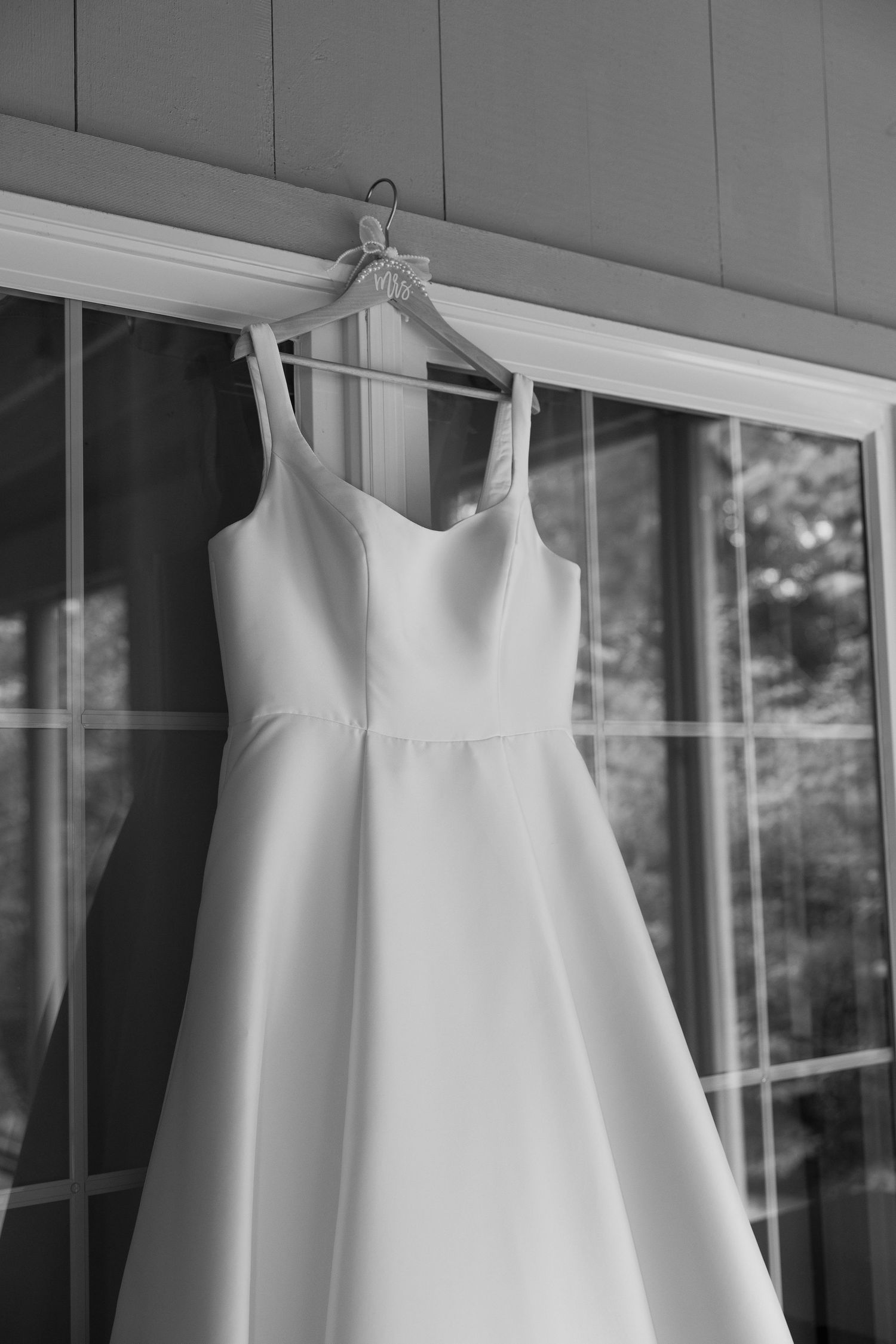Black and white photo of an elegant A-line wedding dress hanging on a hanger in front of large windows.