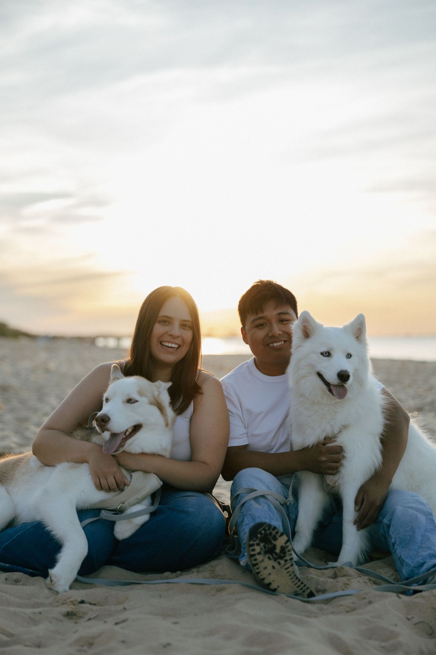 Happy couple sitting on beach at sunset with two white Samoyed dogs, smiling at camera.