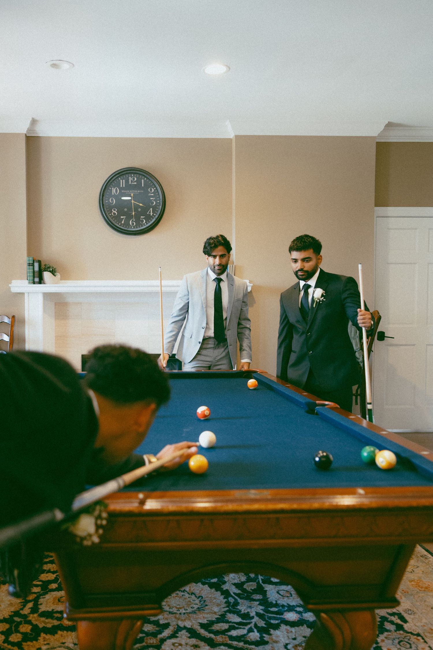 Three men in suits playing pool on a blue felt billiard table in an elegant room with a wall clock.