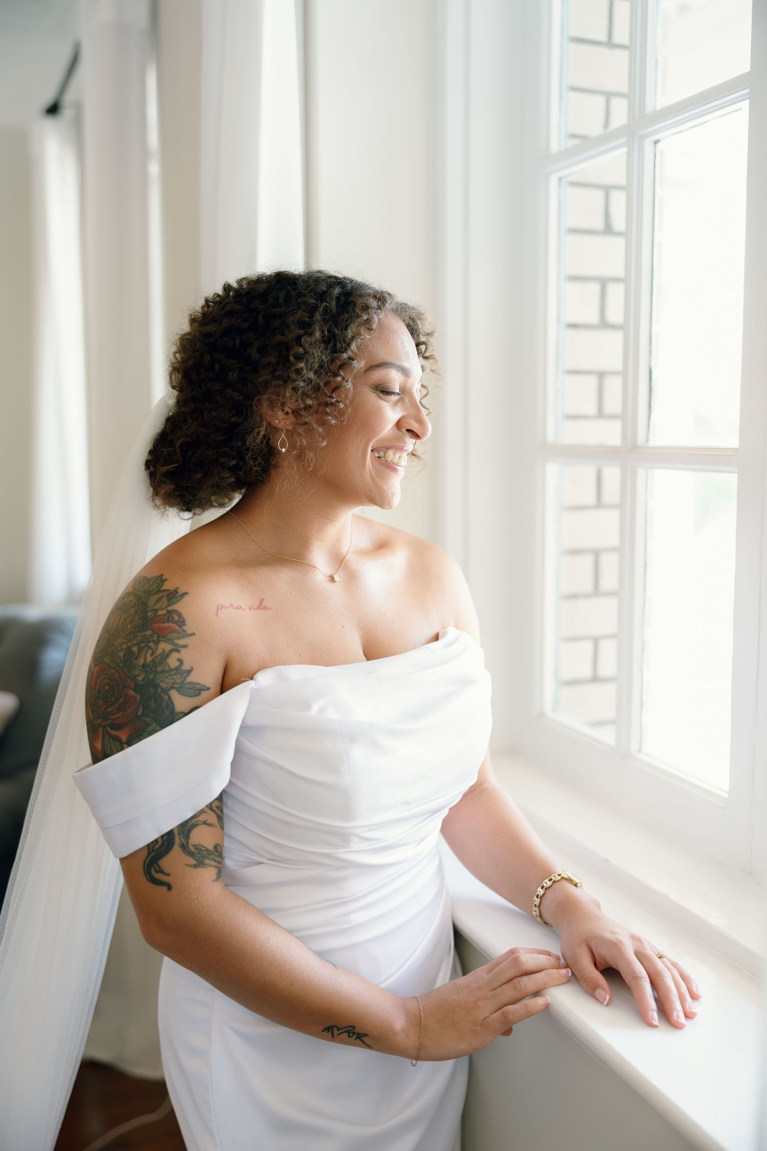 Smiling bride in off-shoulder white gown with tattoos gazes out a bright window on her wedding day.