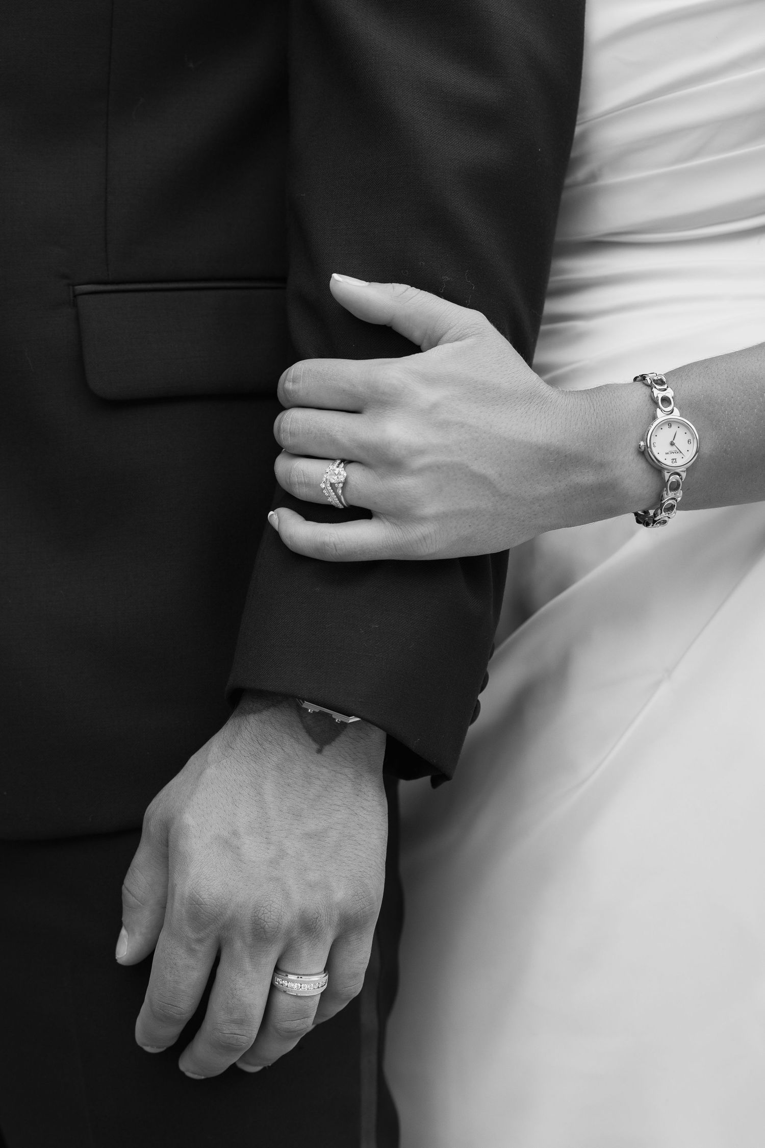 Black and white photo of bride and groom hands showing wedding rings and bracelet watch.