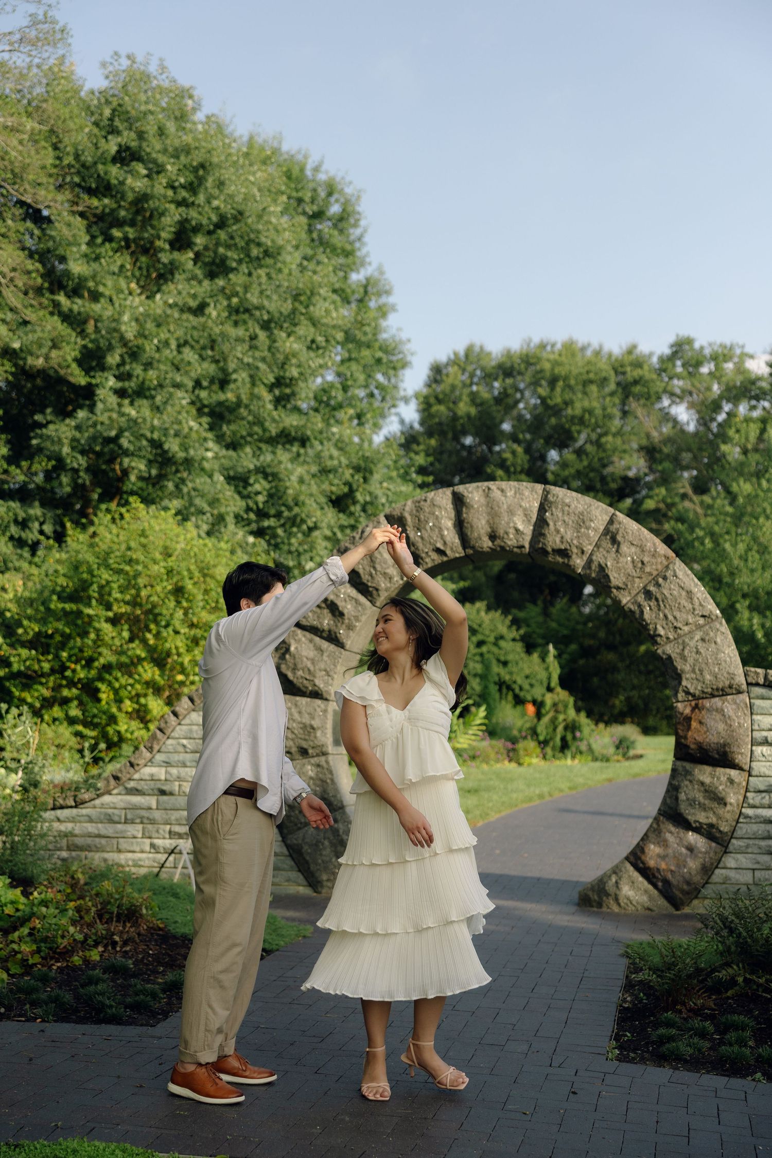 Couple dancing near a circular stone arch in a lush garden, woman in white tiered dress being twirled by man in white shirt.