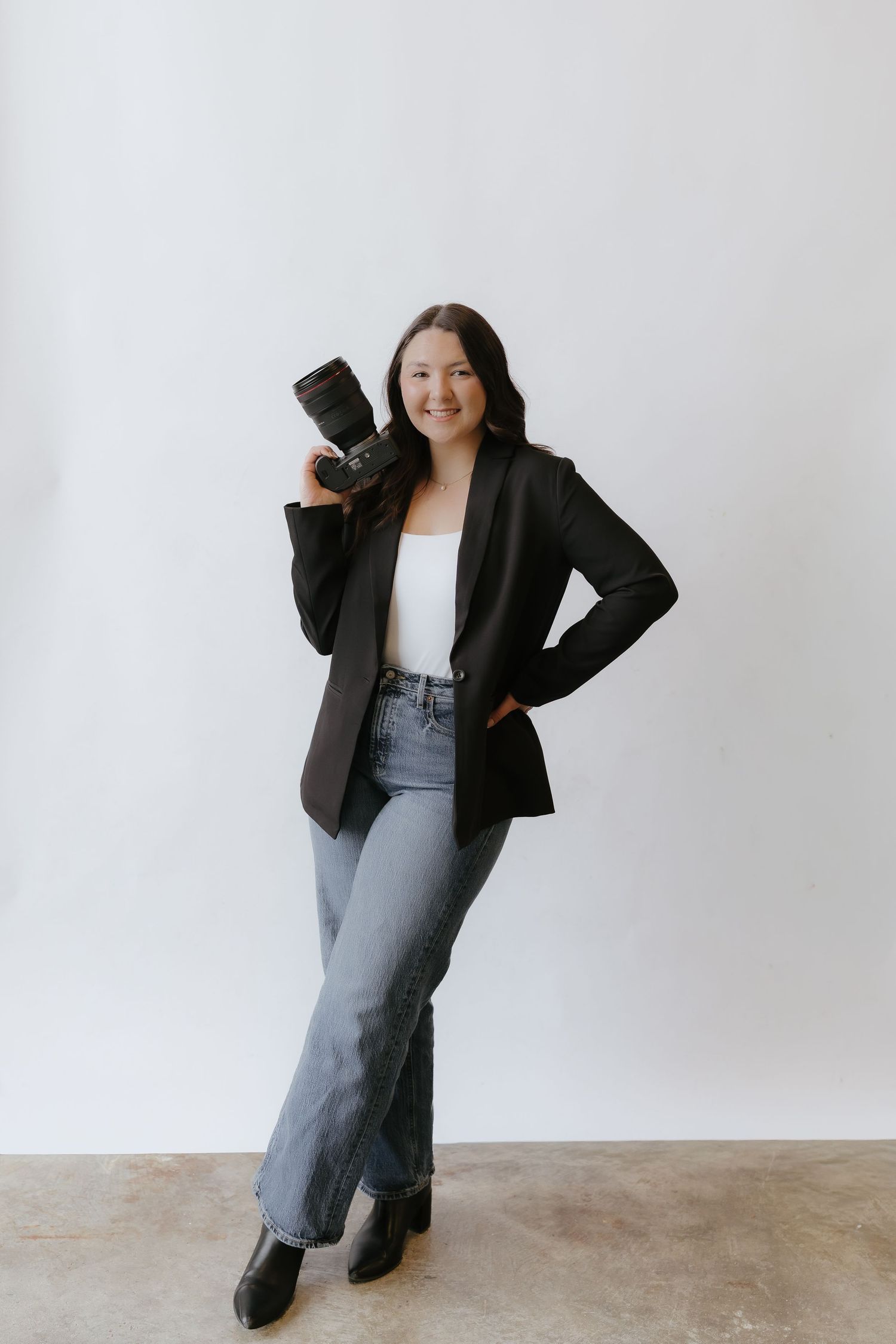 Female photographer in black blazer and jeans posing with Canon camera on shoulder in white studio.