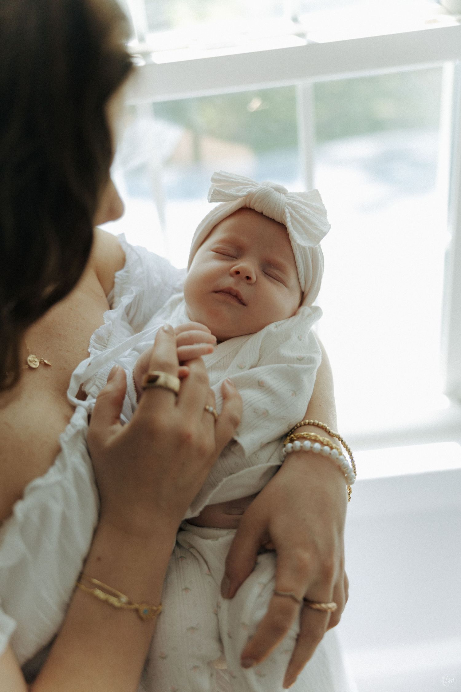 Mother in white dress cradling sleeping newborn baby wearing white bow headband near bright window.