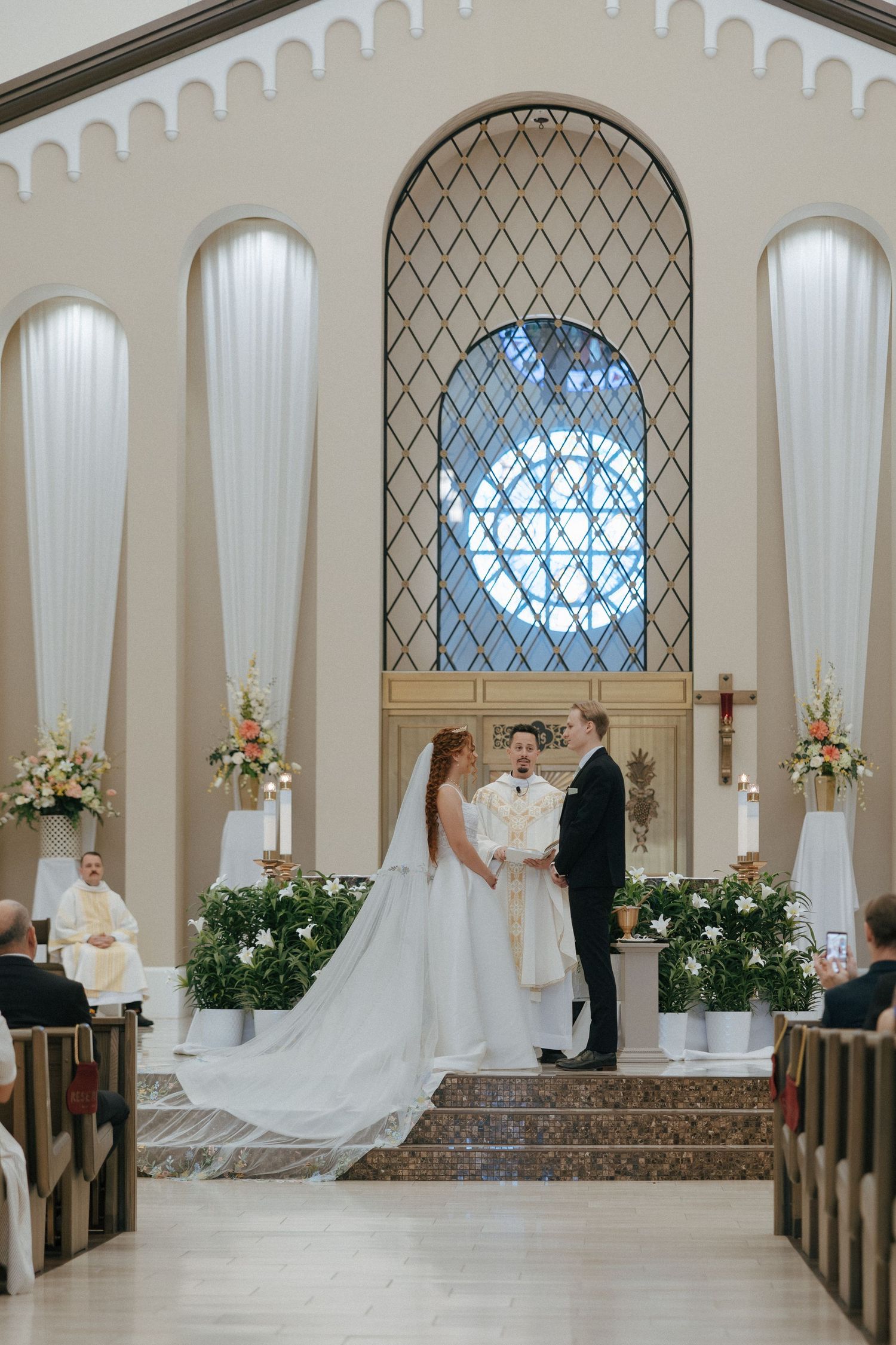 Bride and groom exchanging vows at elegant church altar with arched stained glass window and floral arrangements.
