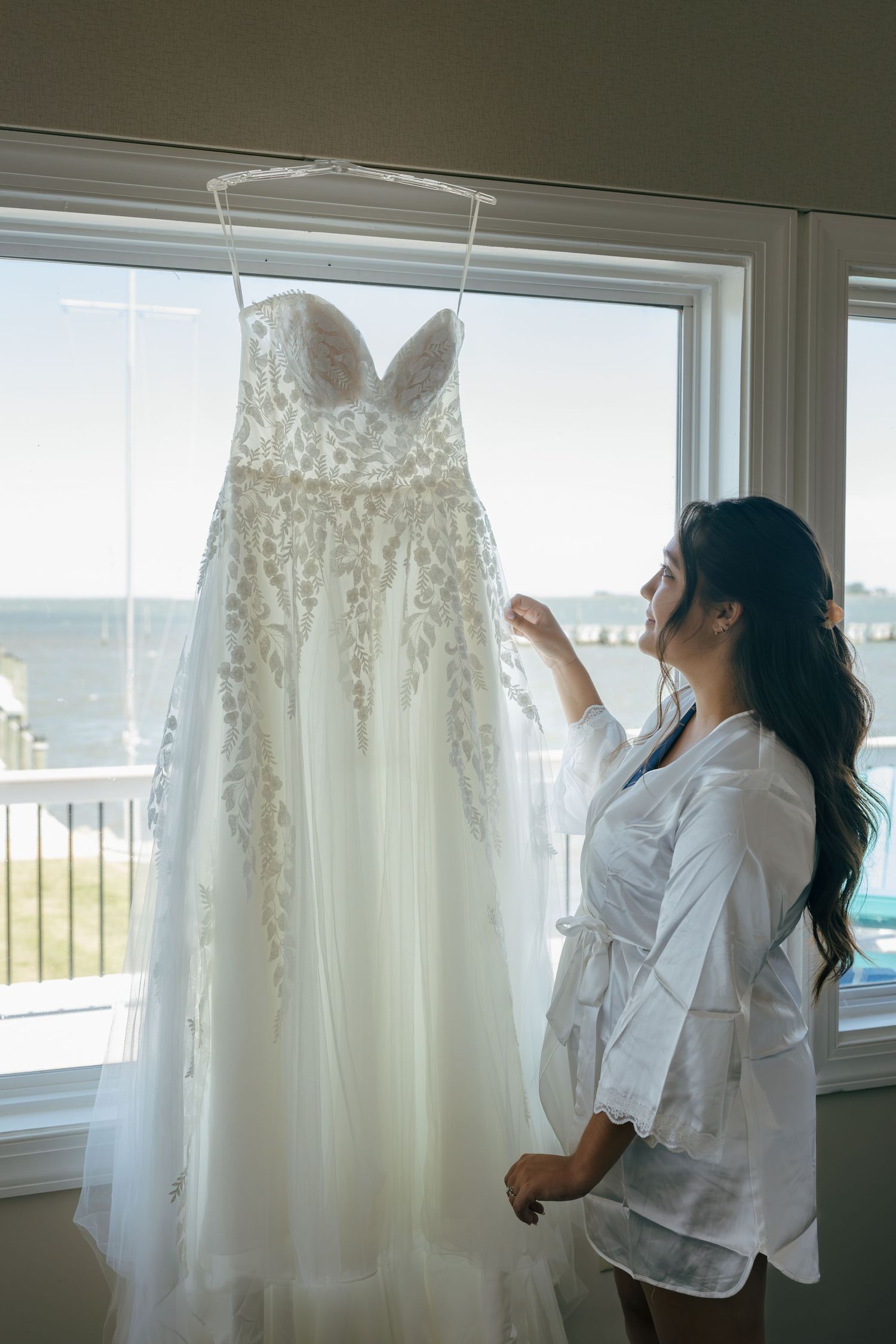 Bride in white robe admiring her lace wedding gown hanging by a window with ocean views.