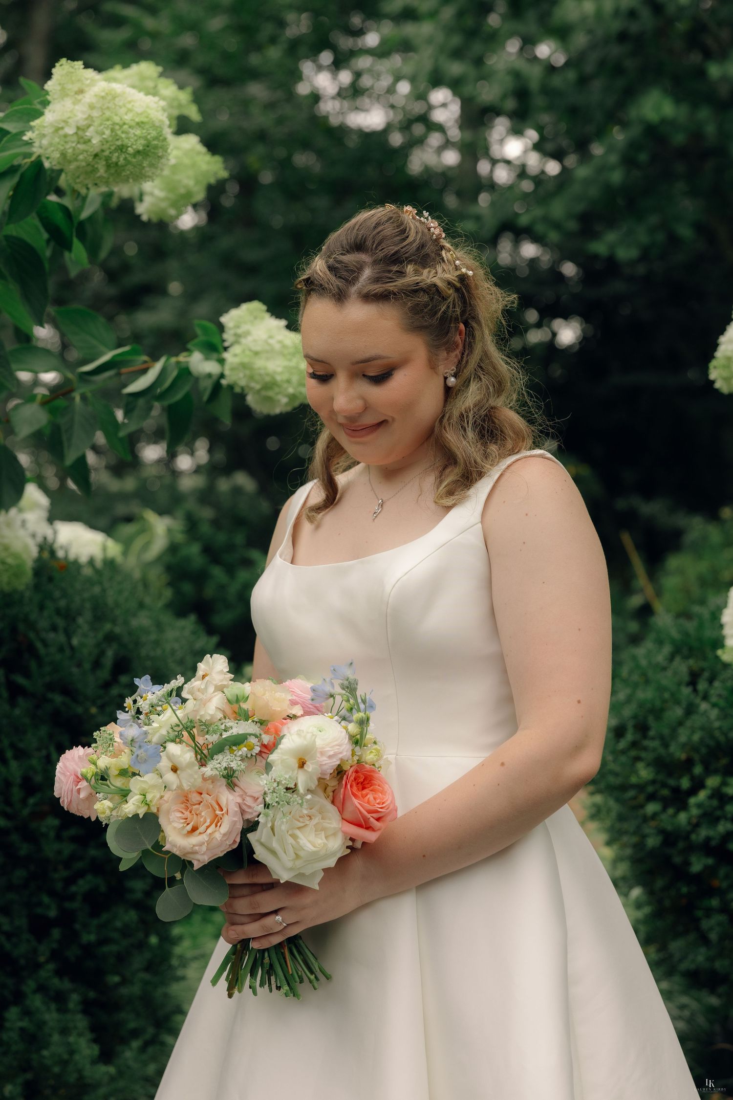 Bride in white gown holding pastel bouquet of roses and blue flowers, smiling in lush garden with hydrangeas.