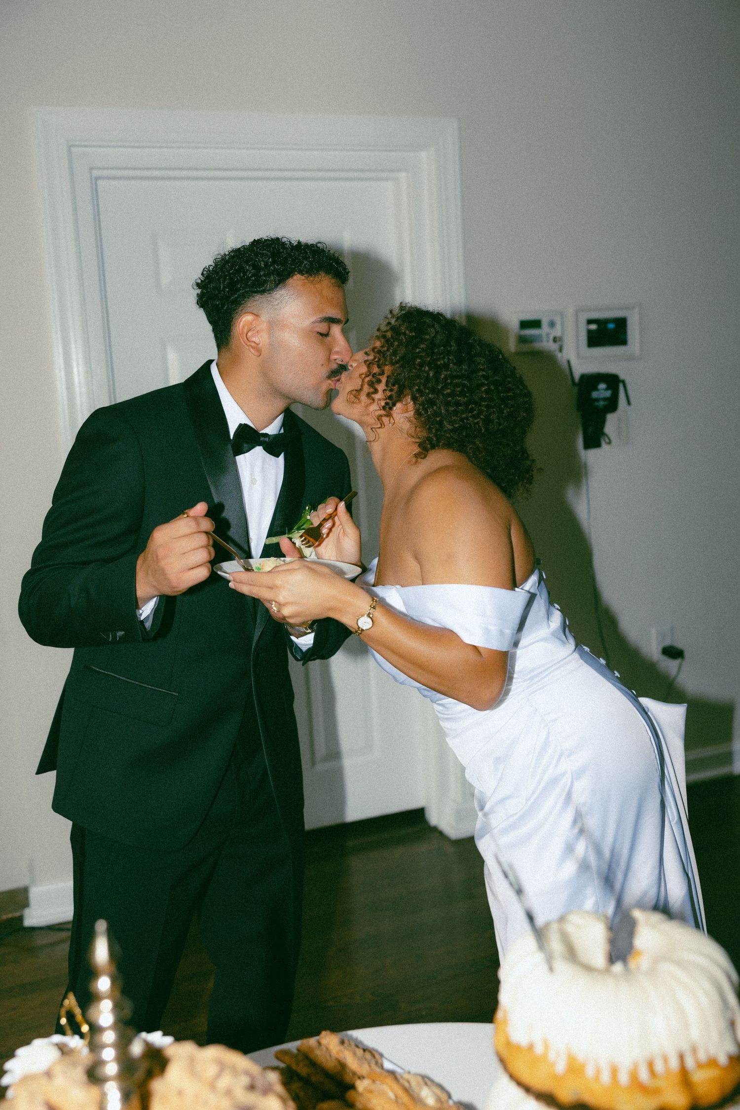Bride and groom sharing a kiss while cutting their wedding cake, dressed in elegant white gown and black tuxedo.