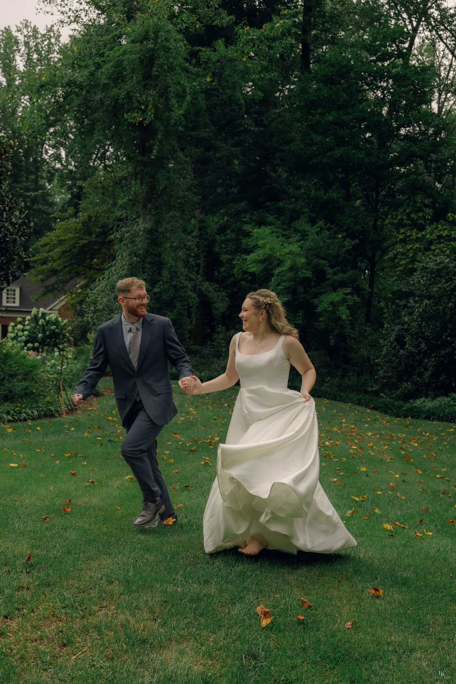 A joyful bride in a white gown and groom in a gray suit hold hands while running across a lush green lawn.