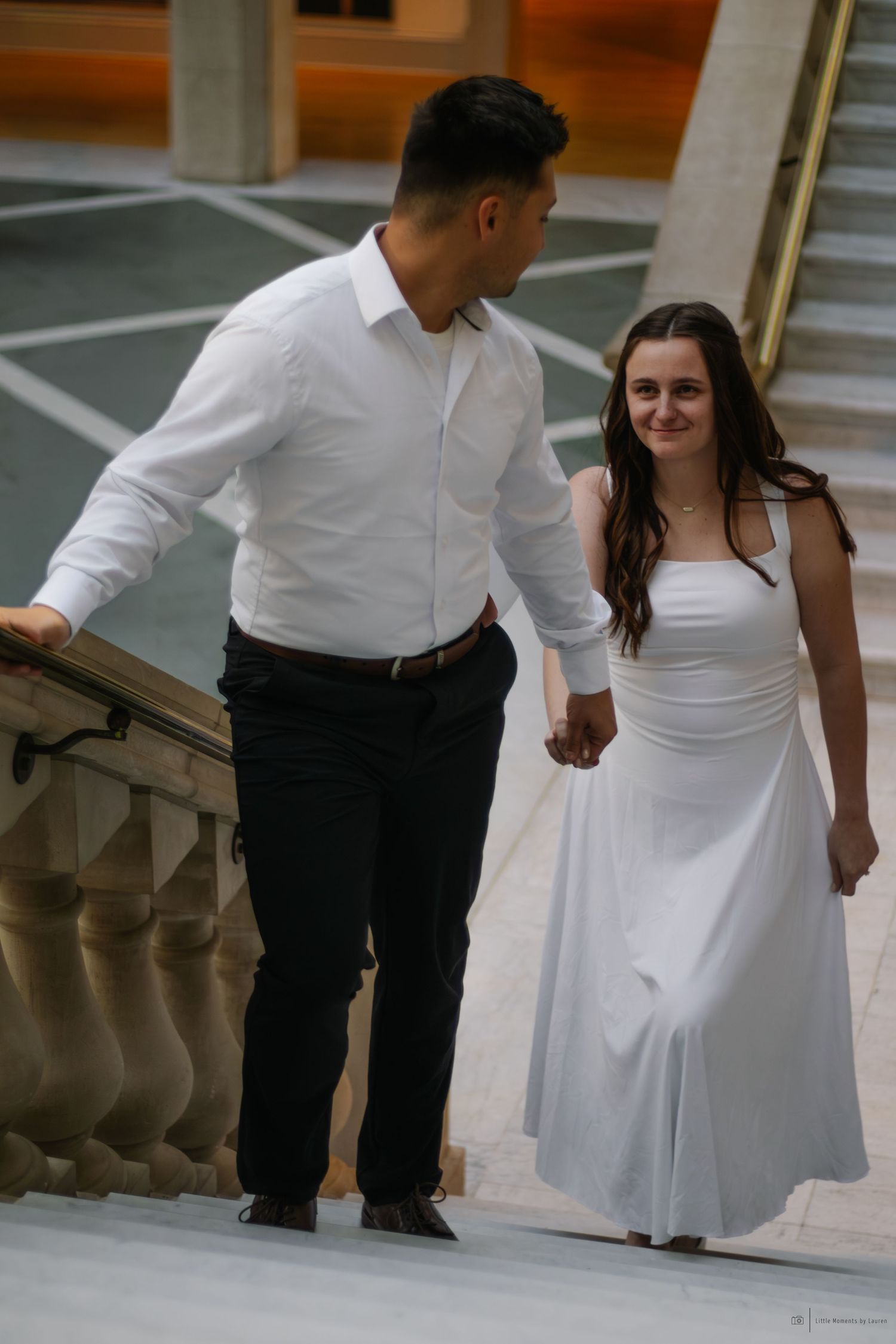 Couple in white outfits holding hands while descending elegant marble stairs in a grand building.