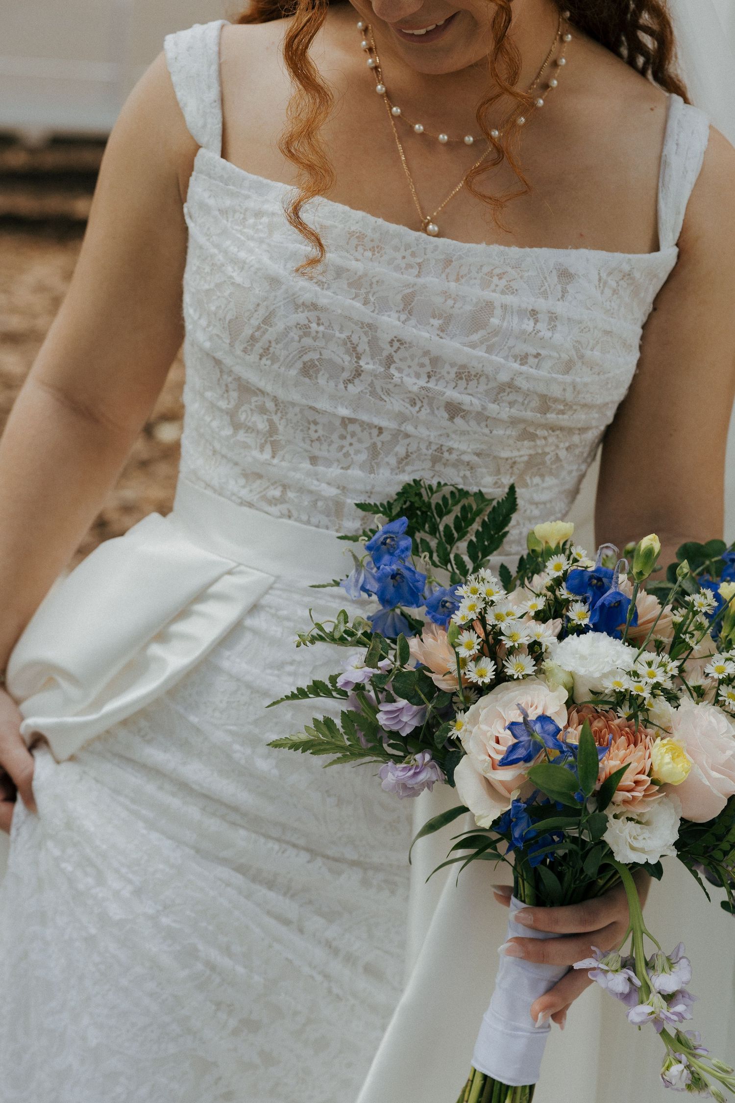 Bride in a square-neck lace wedding gown holding a colorful bouquet of blue, white, and peach flowers.