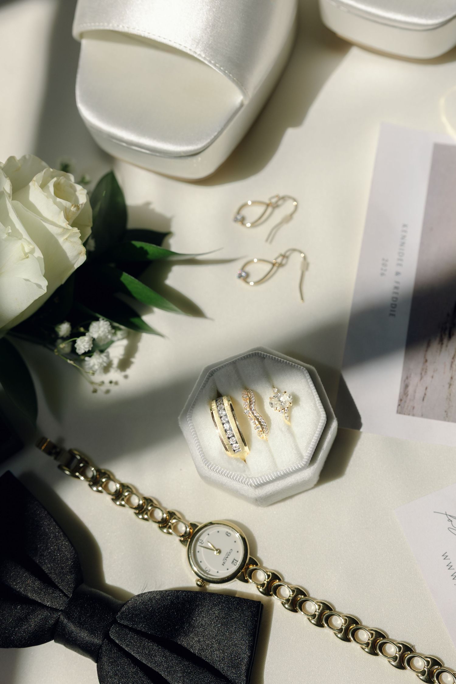 Elegant bridal flat lay with gold rings, pearl watch, white heels, bow tie, rose, and drop earrings on white surface.