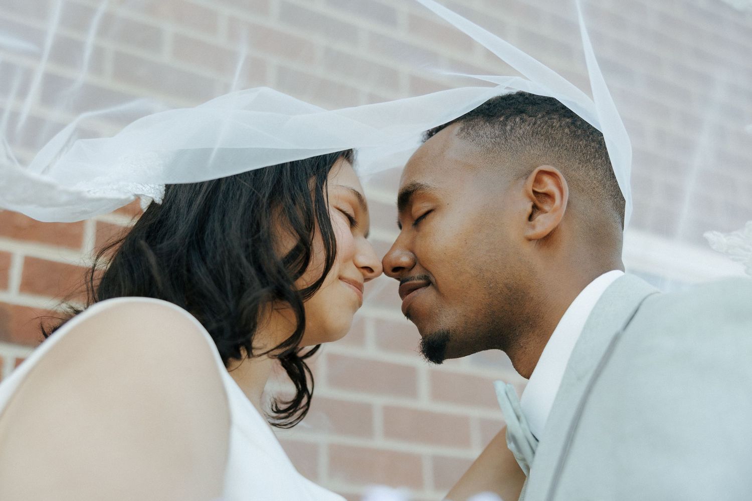 Bride and groom sharing a romantic nose-to-nose moment under a flowing white wedding veil.