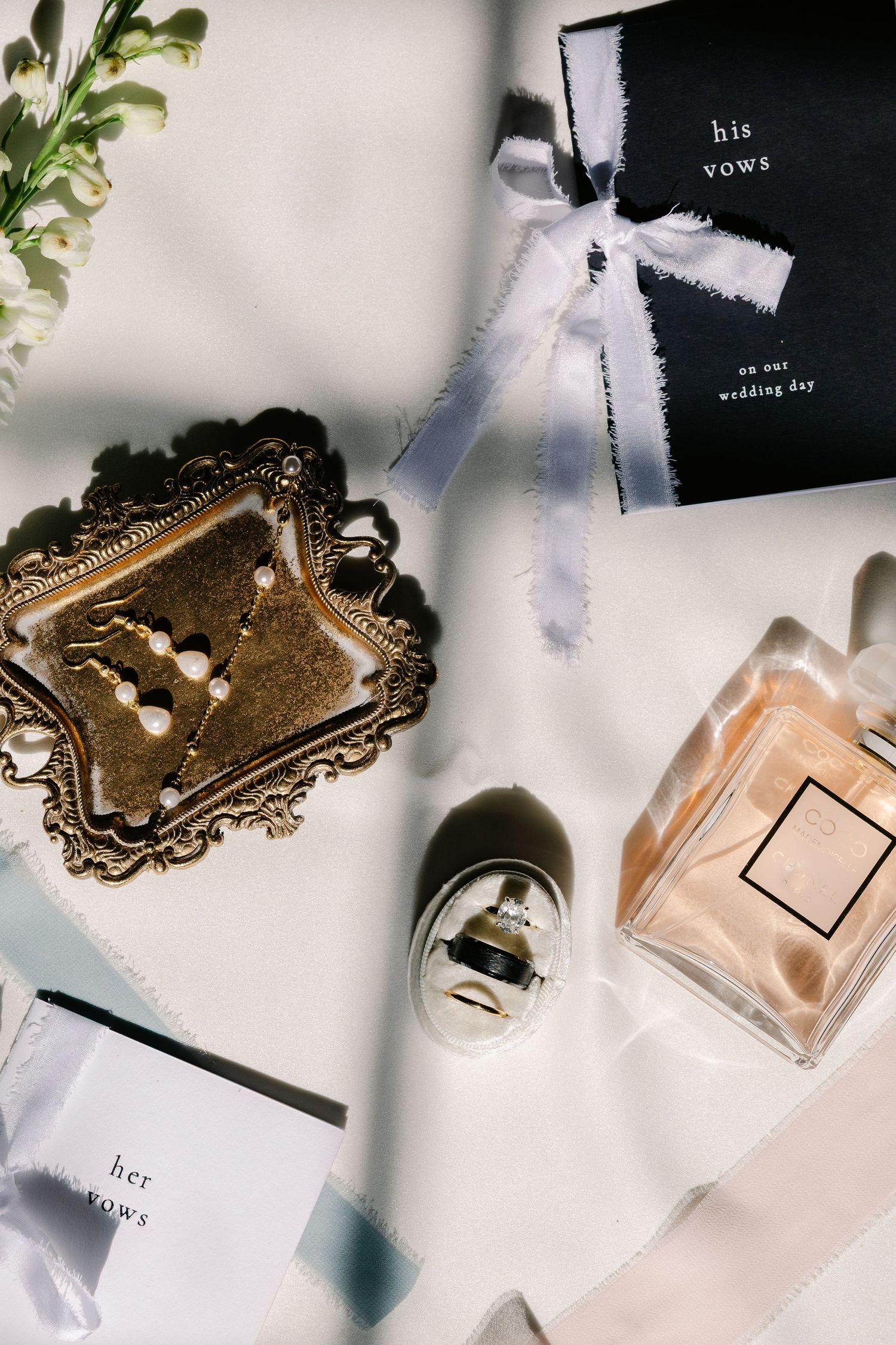 Wedding flat lay with vow books, pearl jewelry, Chanel perfume, and ribbon on white background.