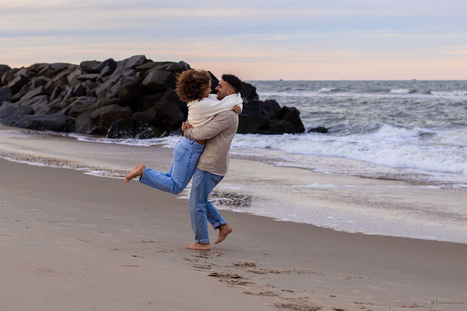 Couple embracing joyfully on a sandy beach at dusk, with rocky jetty and ocean waves in the background.