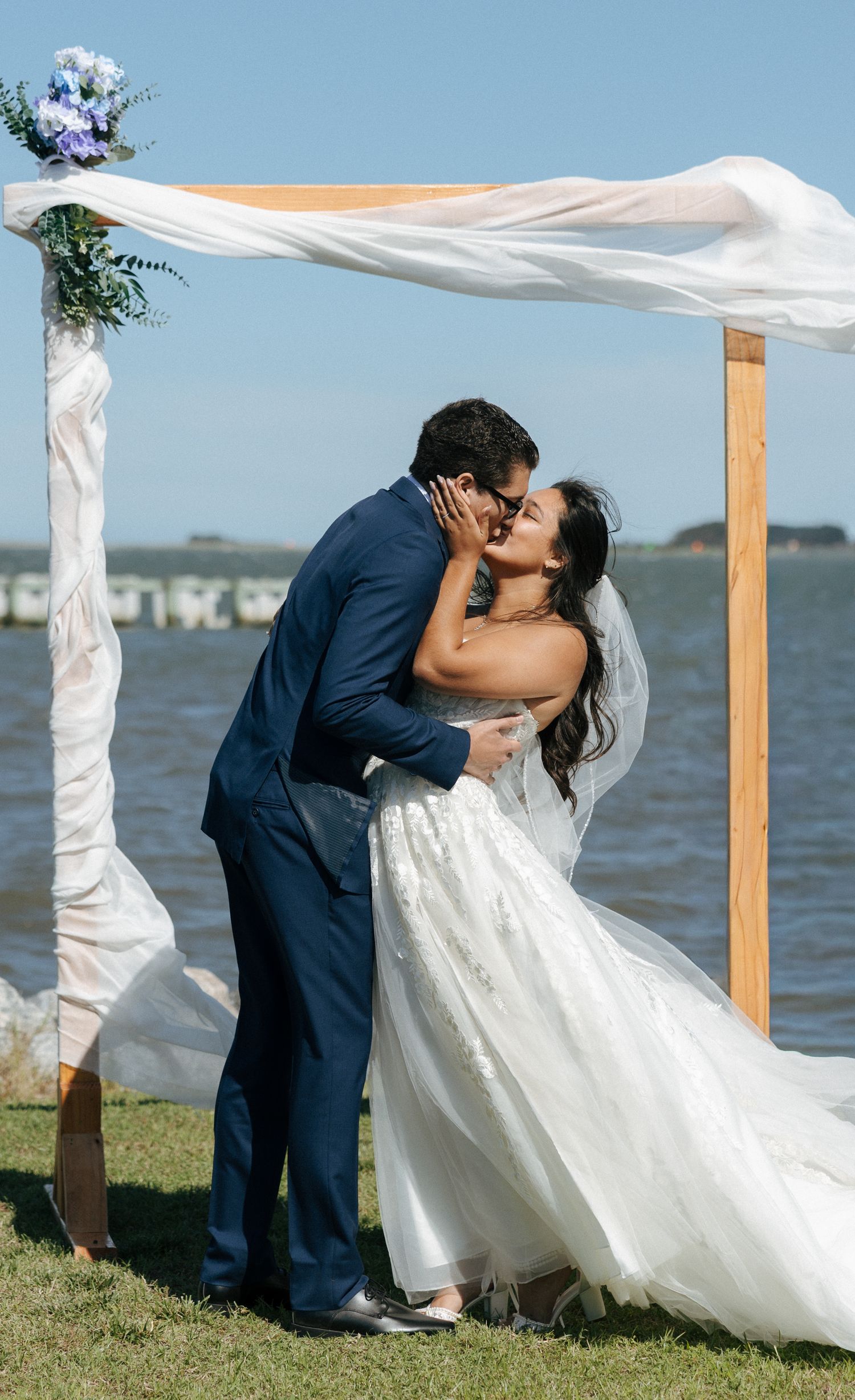 Bride and groom sharing first kiss under wooden arch draped with white fabric beside waterfront on sunny day.