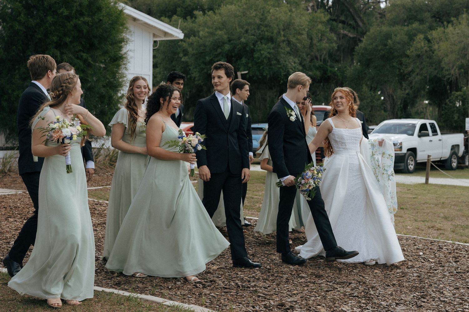 Wedding party in sage green bridesmaid dresses with bride in white gown and groom in black suit outdoors.