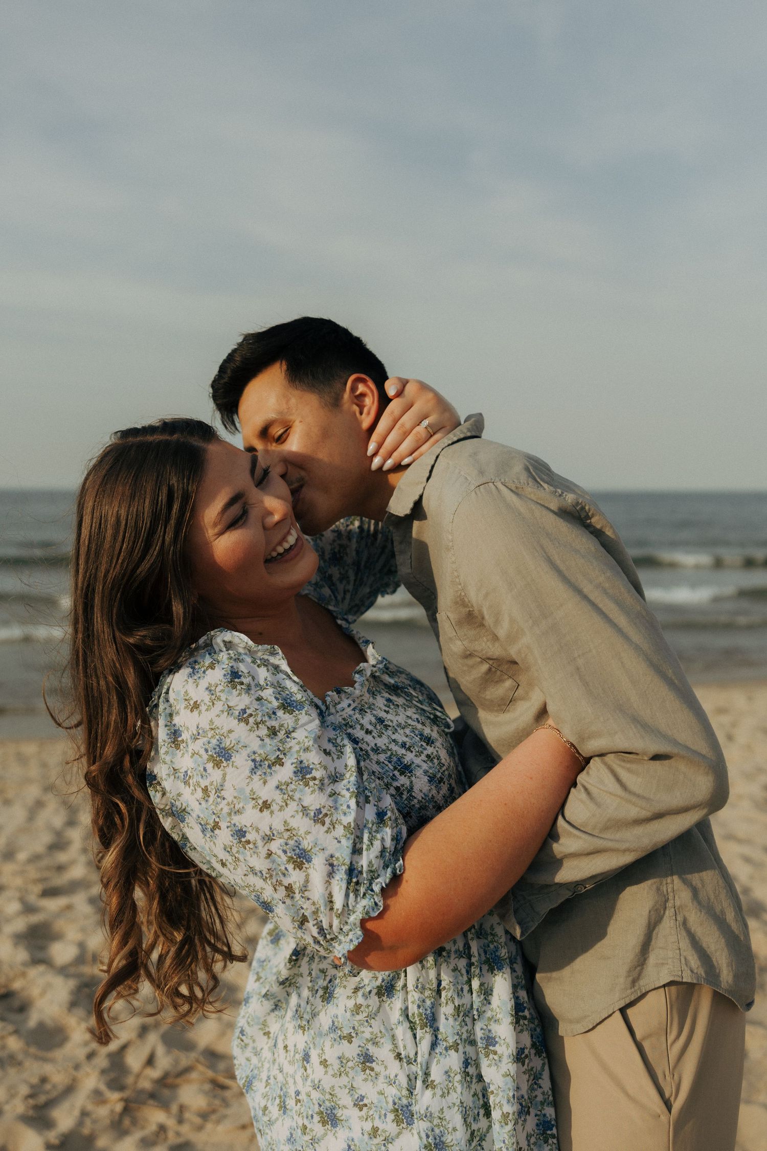 Couple embracing on a sandy beach at sunset, man kissing woman's cheek as she smiles joyfully.