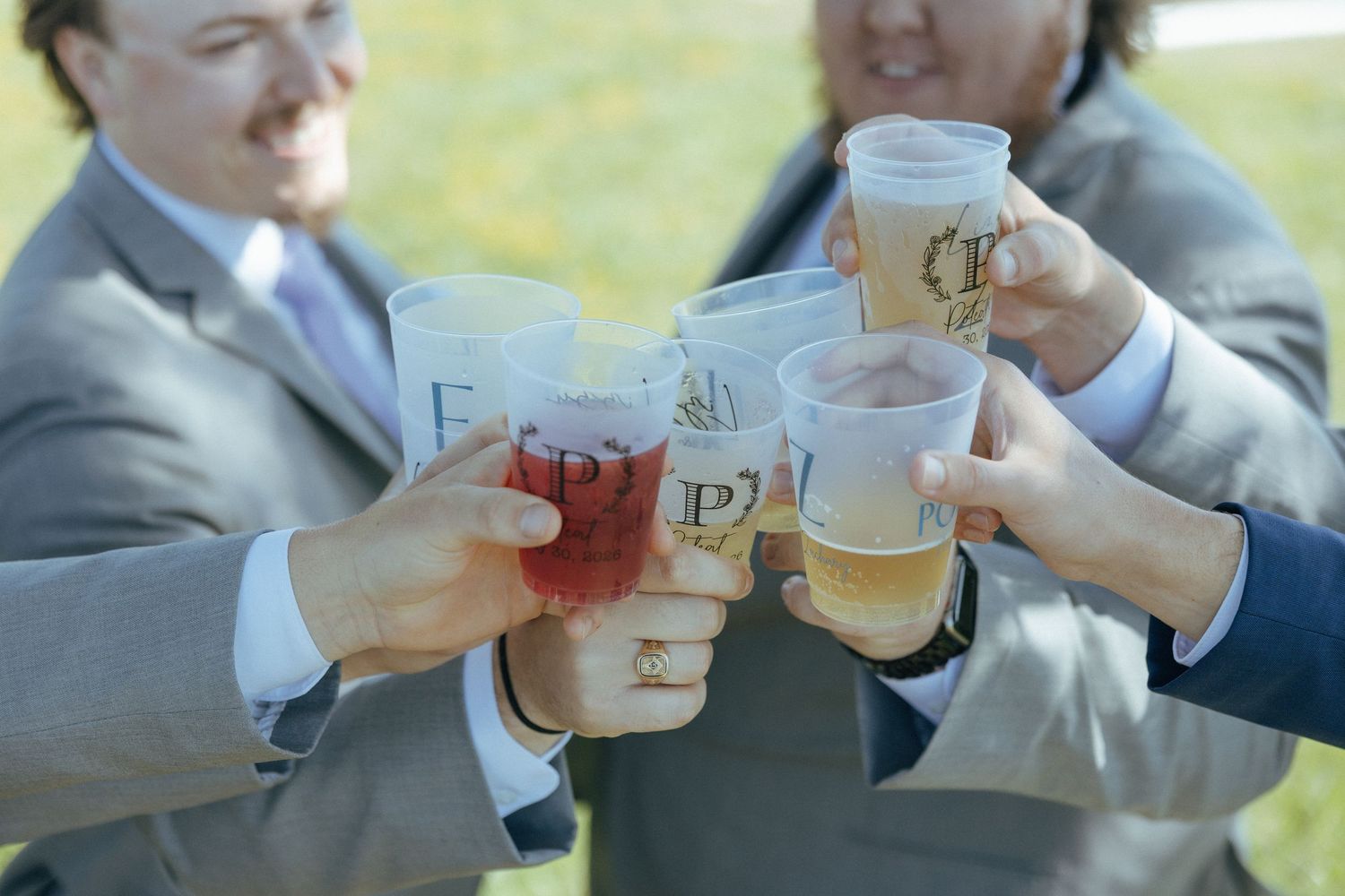 Group of men in suits toasting with personalized plastic cups filled with beer and red drinks outdoors.