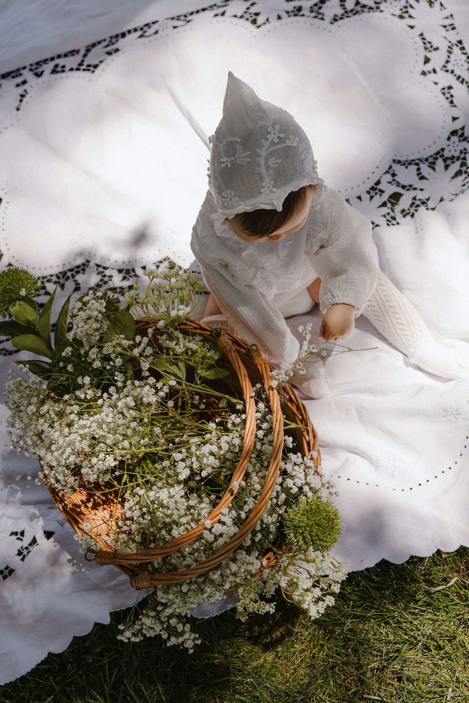 Baby in white christening gown and bonnet reaching into a wicker basket of white wildflowers.