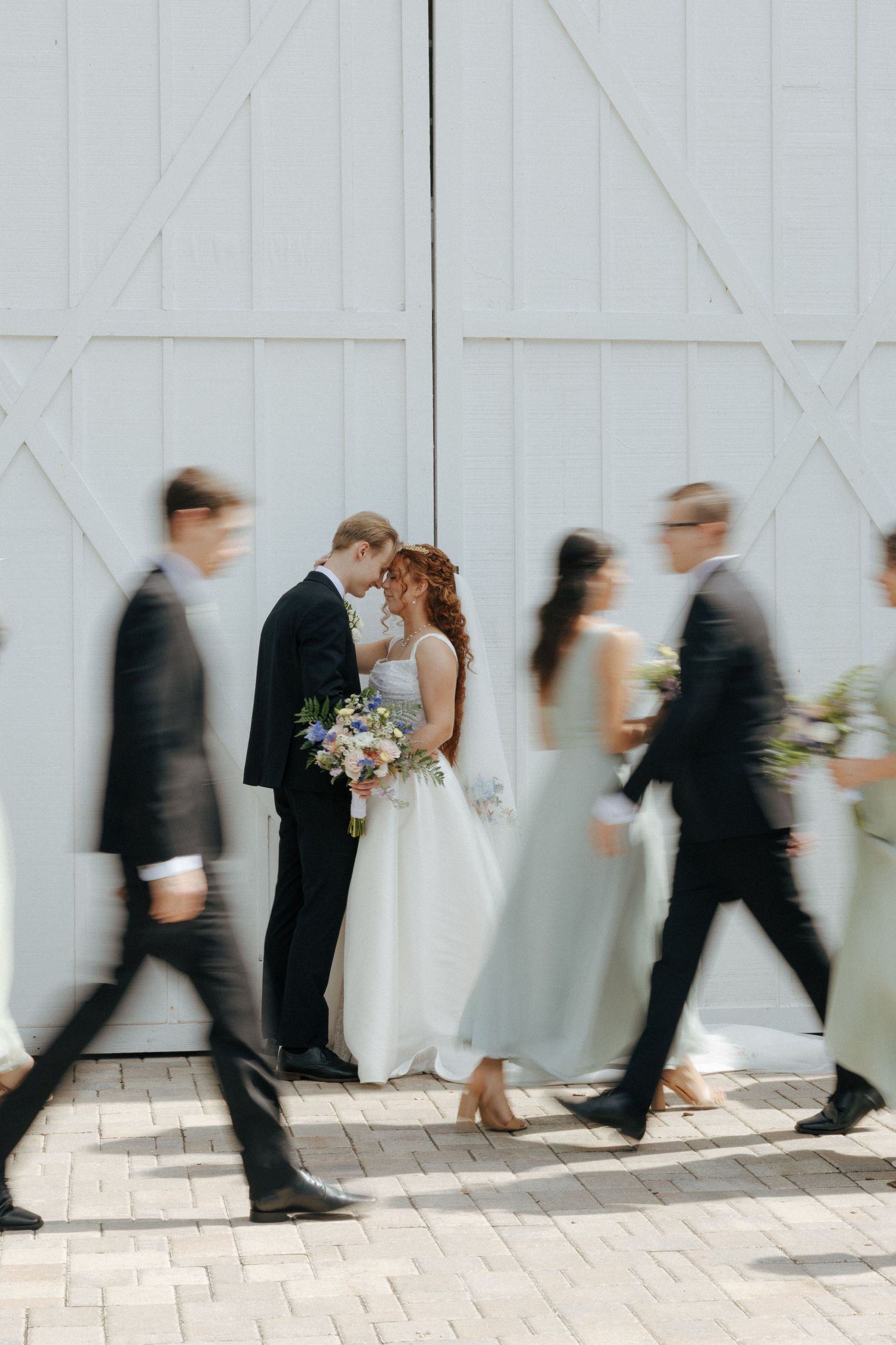Bride and groom share a tender moment while wedding party walks past in motion blur near white barn doors.