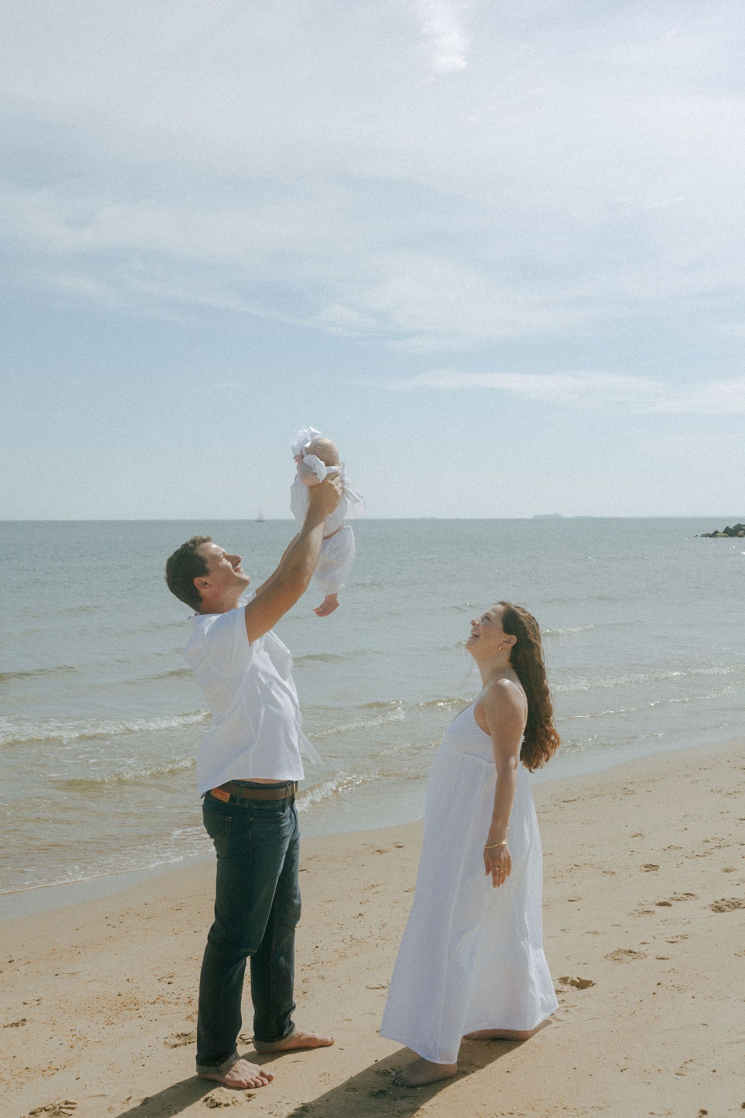 Father lifts baby in the air while mother watches joyfully on a sunny sandy beach.